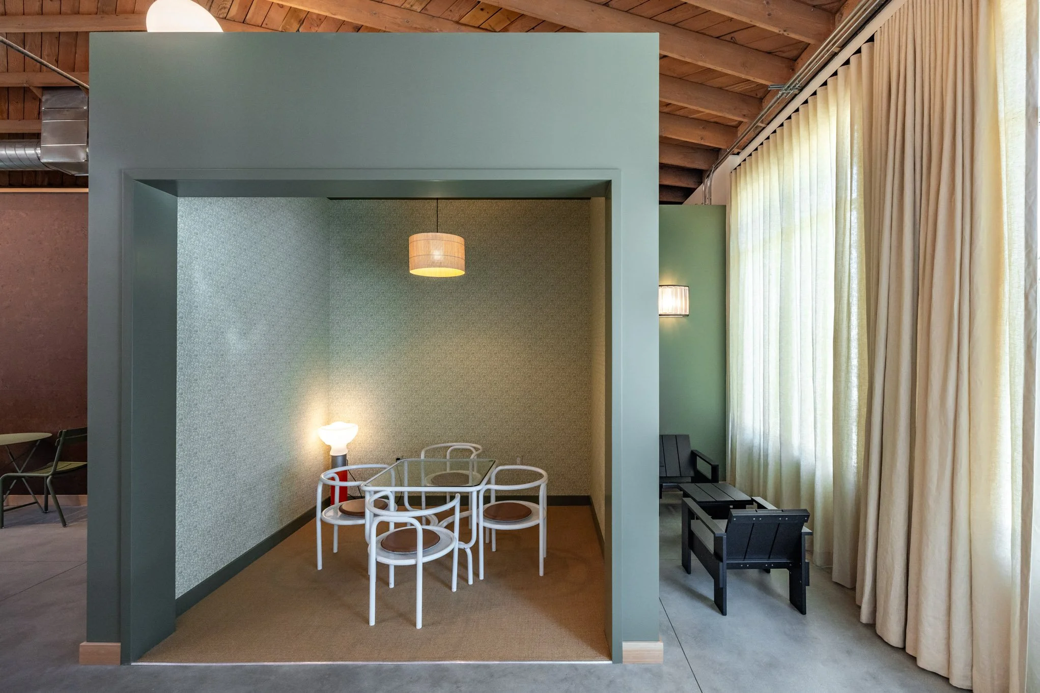 A cozy vintage-style corner with a small round white table and four matching white chairs with brown seats, illuminated by a small standing lamp, inside a pastel-colored nook with textured wallpaper, adjacent to a seating area with black chairs and l