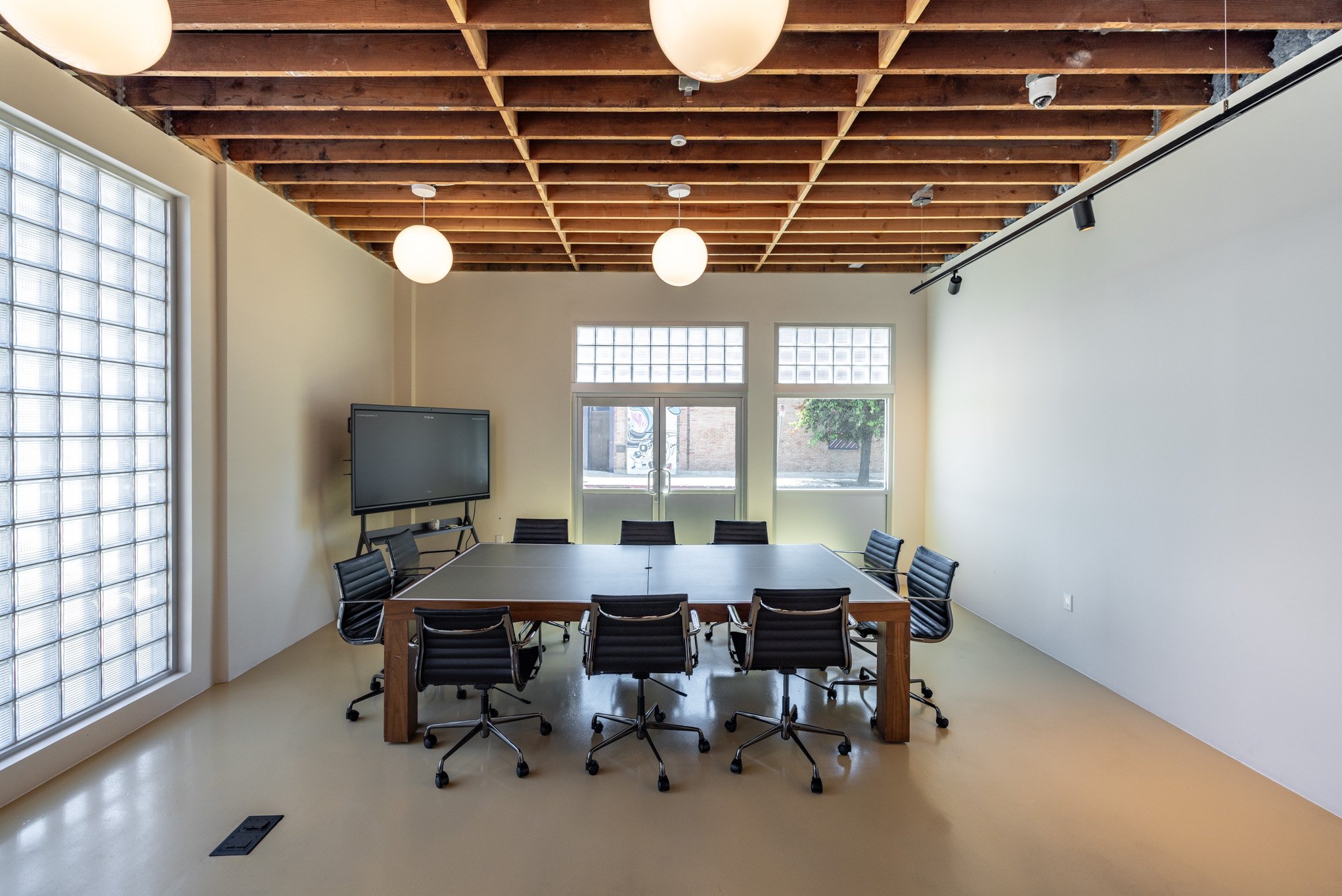Empty modern conference room with a long rectangular table, eight black swivel chairs, a large flat-screen TV on a stand, large windows, and a glass door, with exposed wooden ceiling beams and round ceiling lights.