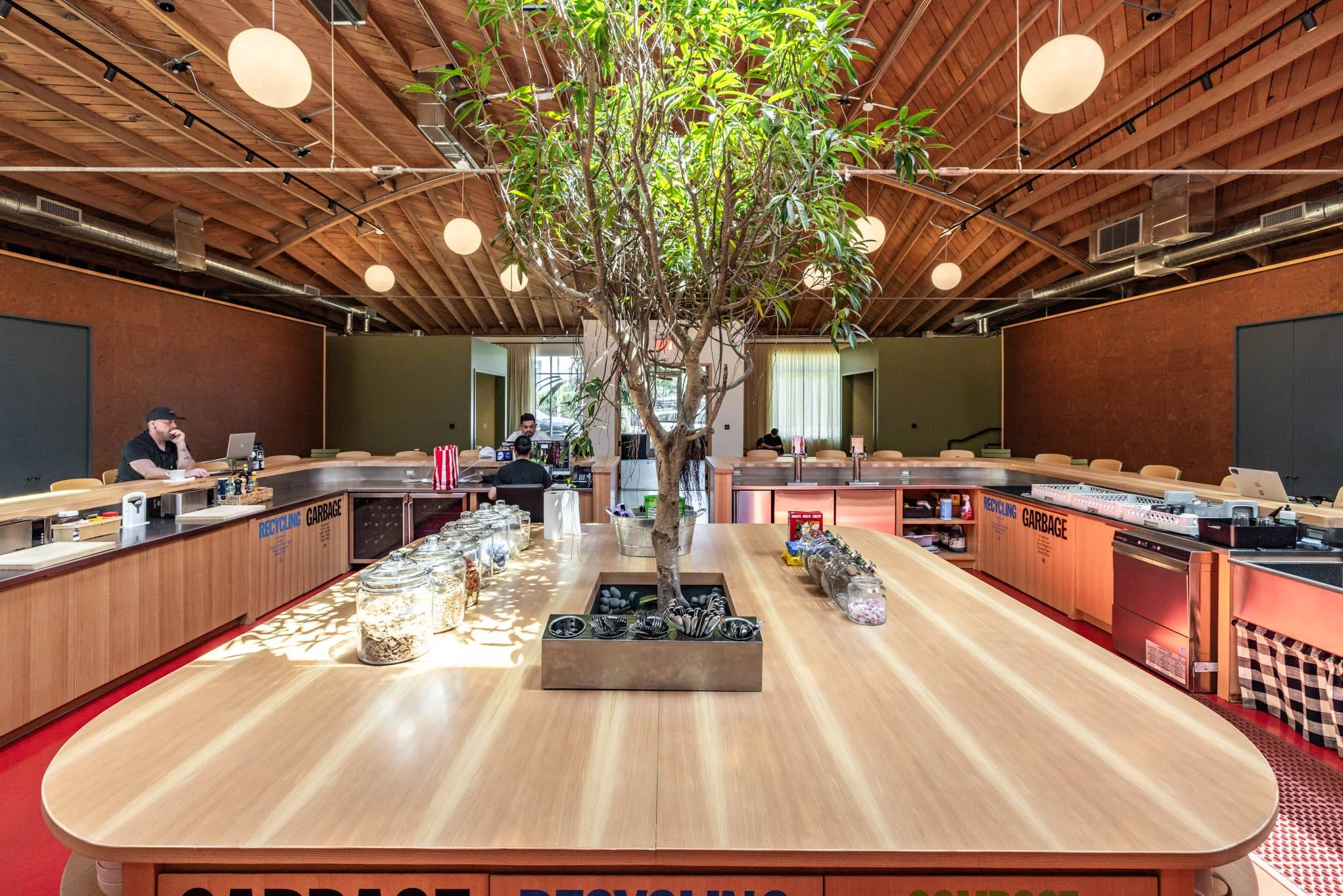 Indoor space with a large wooden table in the center, a tall tree growing from the middle, and a wooden ceiling with round hanging lights. There are people working at counters with catering and recycling signs, and a bright window in the background.
