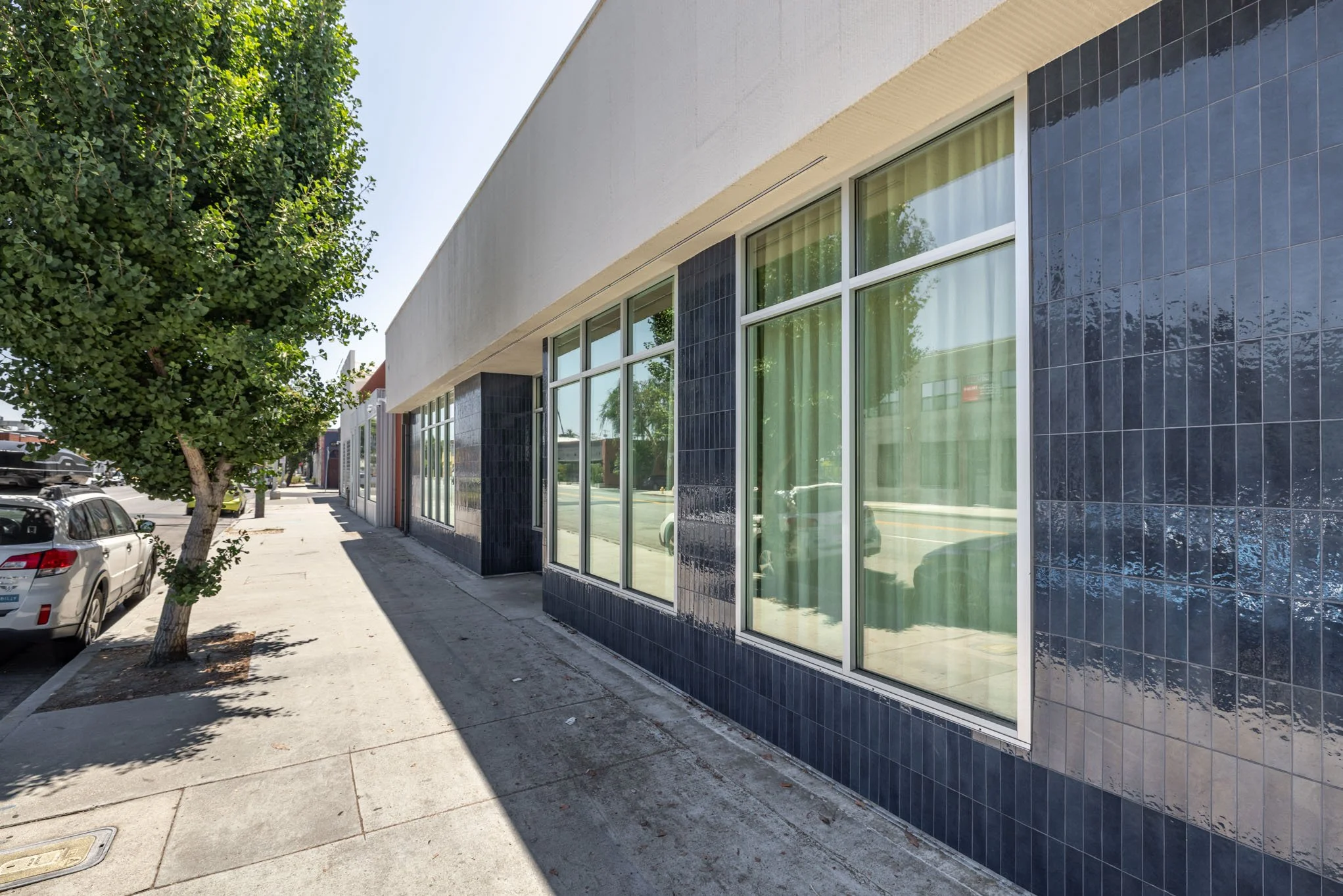 Modern building with large glass windows and dark blue tile accents on a sunny street with trees and parked cars.
