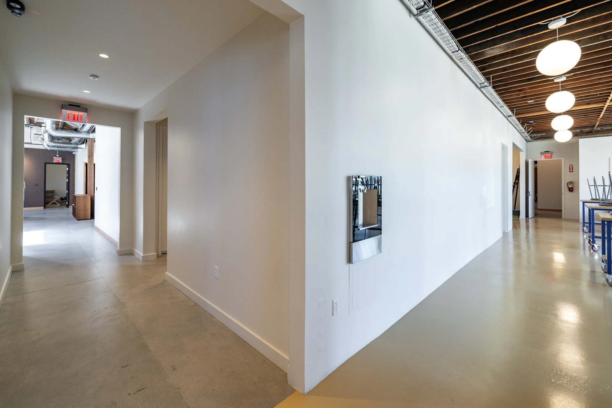 Interior of a modern building with white walls, wooden ceiling with hanging lights, and a polished concrete floor. Visible rooms and hallways, some with furniture stacked, and an exit sign.