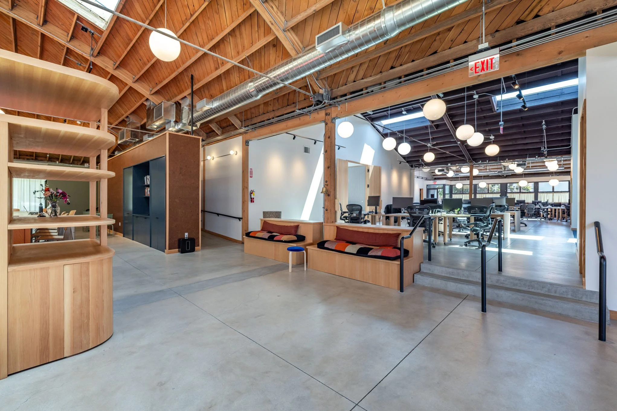 Open office space with modern decor, wooden accents, and large windows allowing natural light, featuring work desks, chairs, and a seating area.