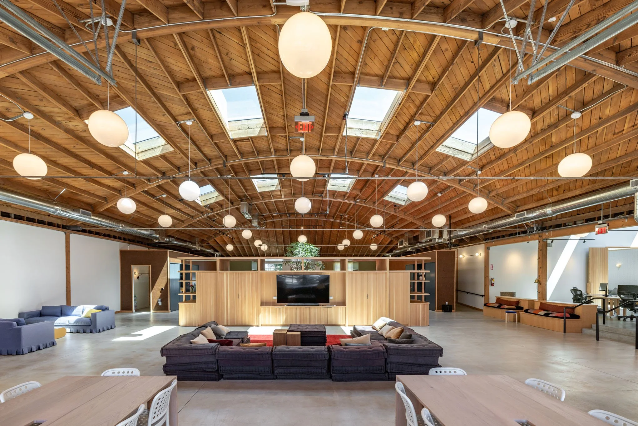 Large open lounge area with a wooden ceiling and skylights, featuring a curved dark sofa with cushions, a large flat-screen TV on a wooden divider, and various seating options including blue couches and built-in benches, with round hanging lights