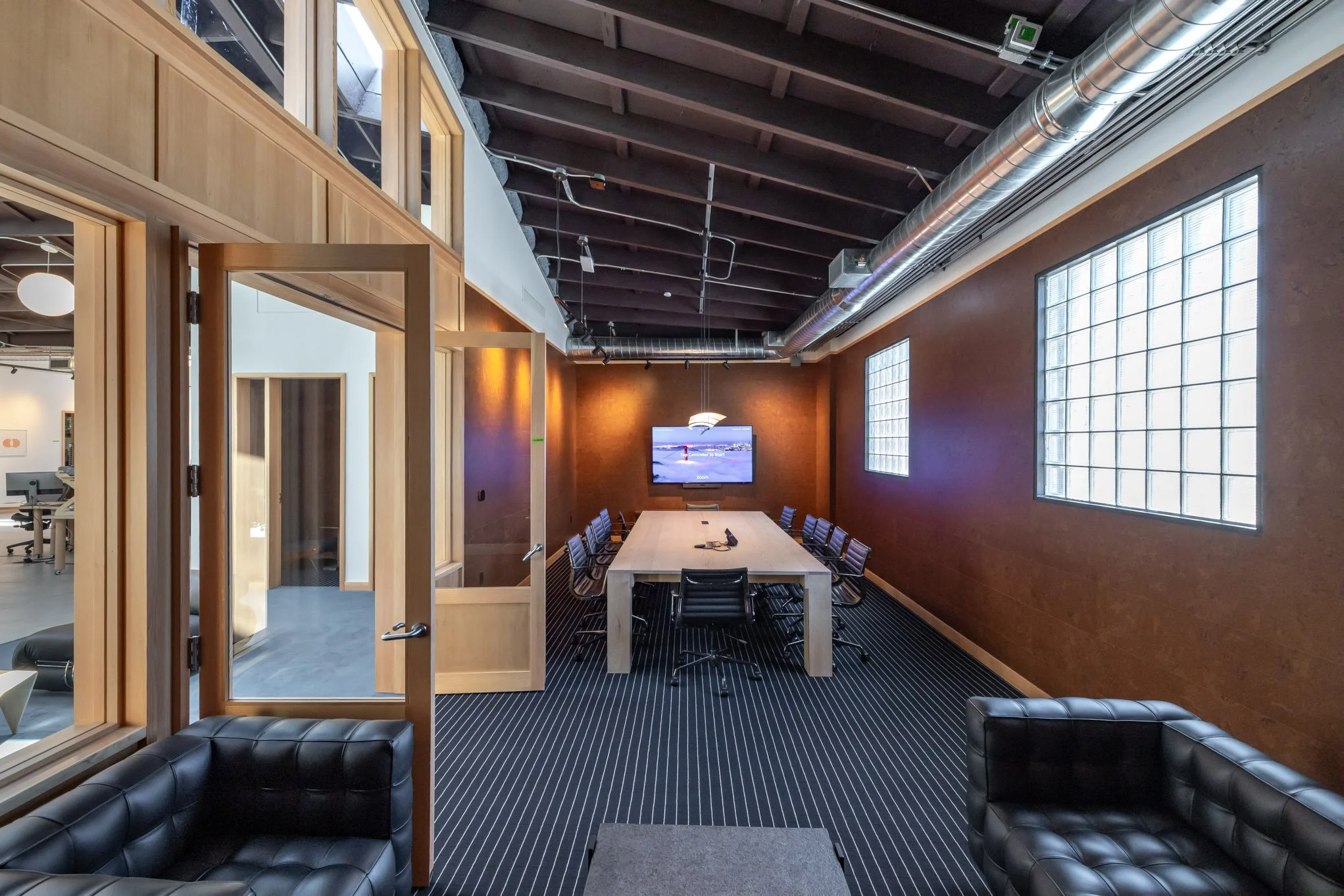 Modern conference room with wooden accents, glass block windows, and a long table with chairs, a large wall-mounted TV at the end, and black leather chairs in the foreground.