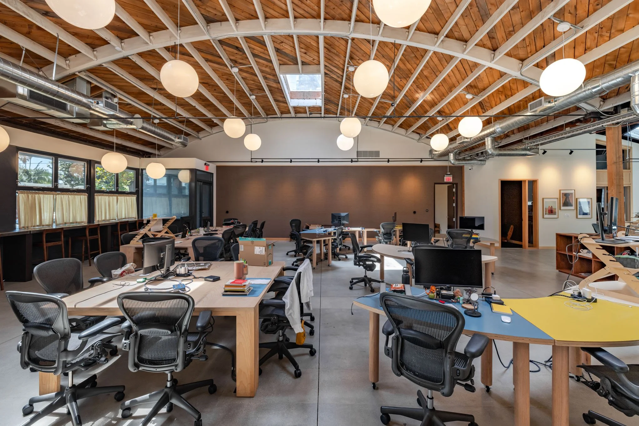 Empty modern office space with multiple workstations, chairs, computers, and large pendant lights hanging from a wooden ceiling.
