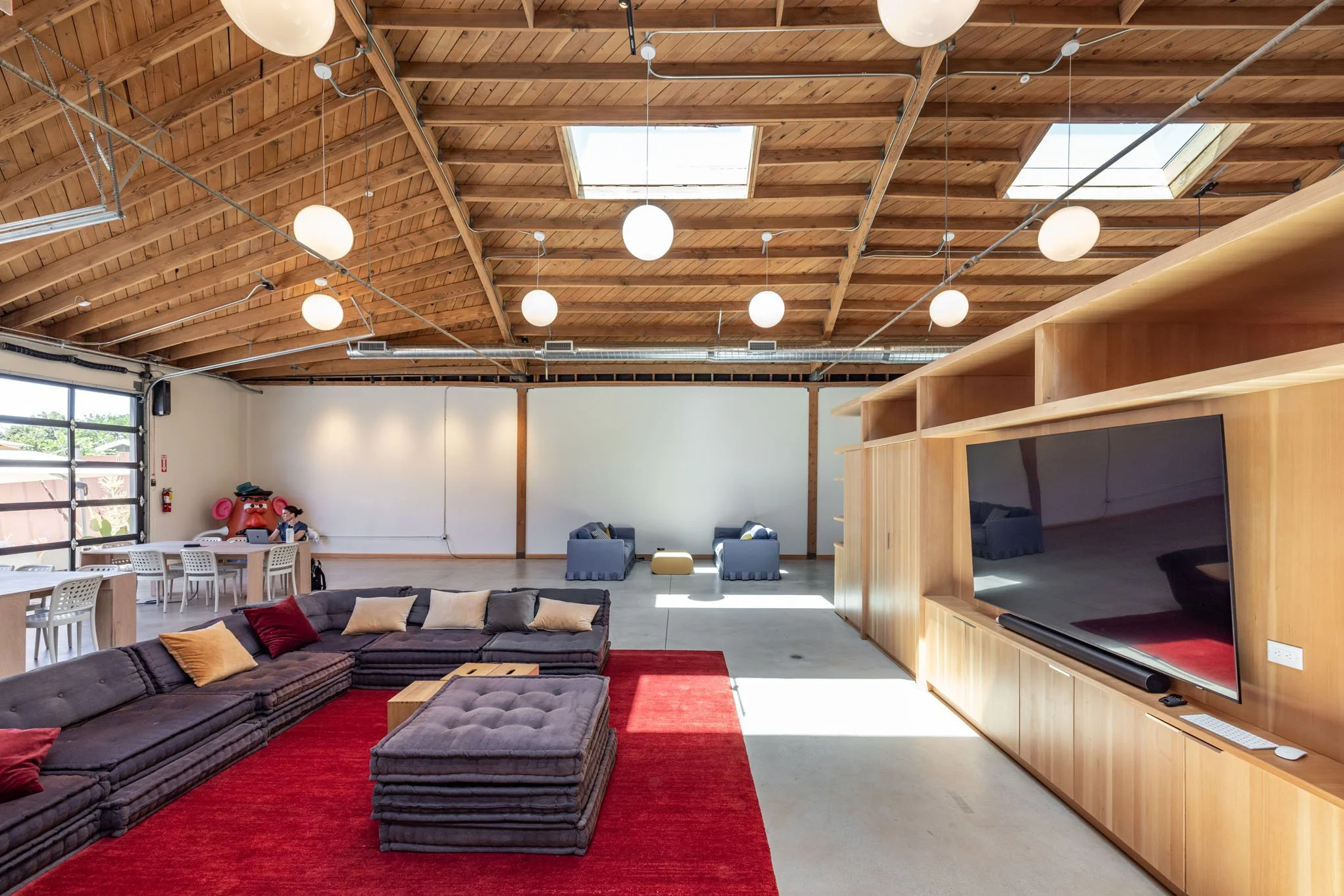 Spacious lounge area with a large sectional sofa on a red rug, a wall-mounted flat screen TV, and a wooden ceiling with skylights and round hanging lights.