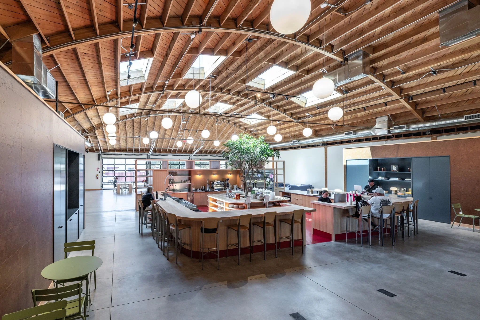 Interior of a modern coffee shop with wooden ceiling and large windows. There is a central island with a tree, seating areas, and customers working on laptops.