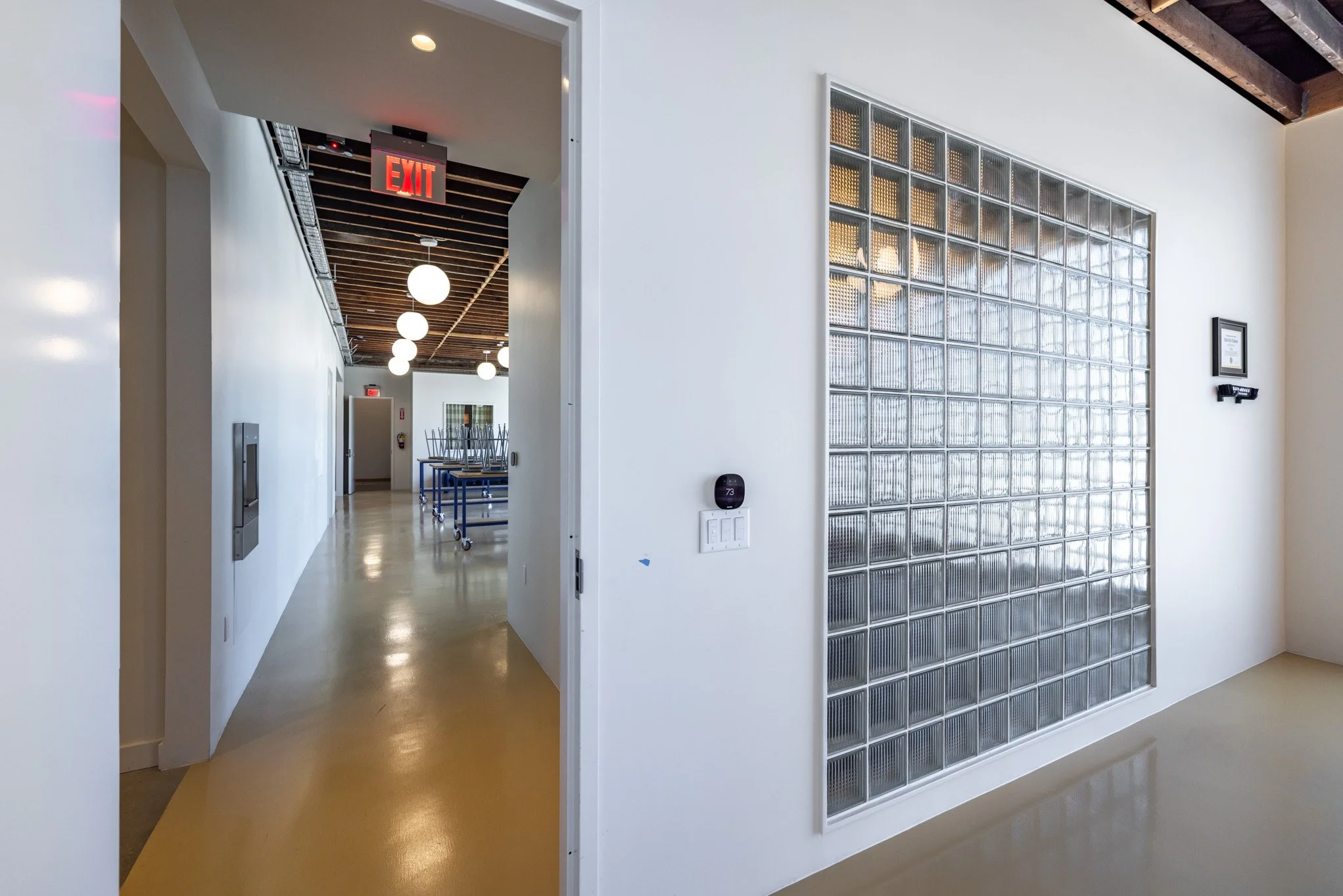 Interior hallway with white walls, glass block wall, and visible ceiling with wooden beams. There are hanging globe lights and a red exit sign hanging from the ceiling. The floor is shiny and light-colored.