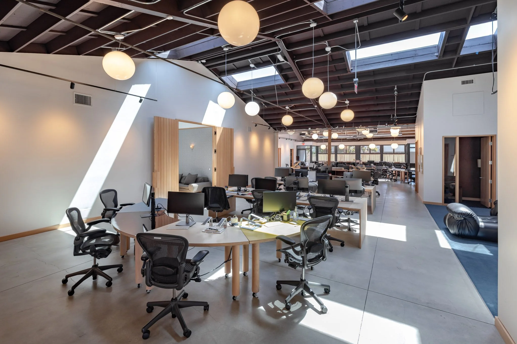 Open-plan modern office with desks, chairs, computers, pendant lights, and a skylight ceiling, featuring natural sunlight and a relaxed workspace atmosphere.