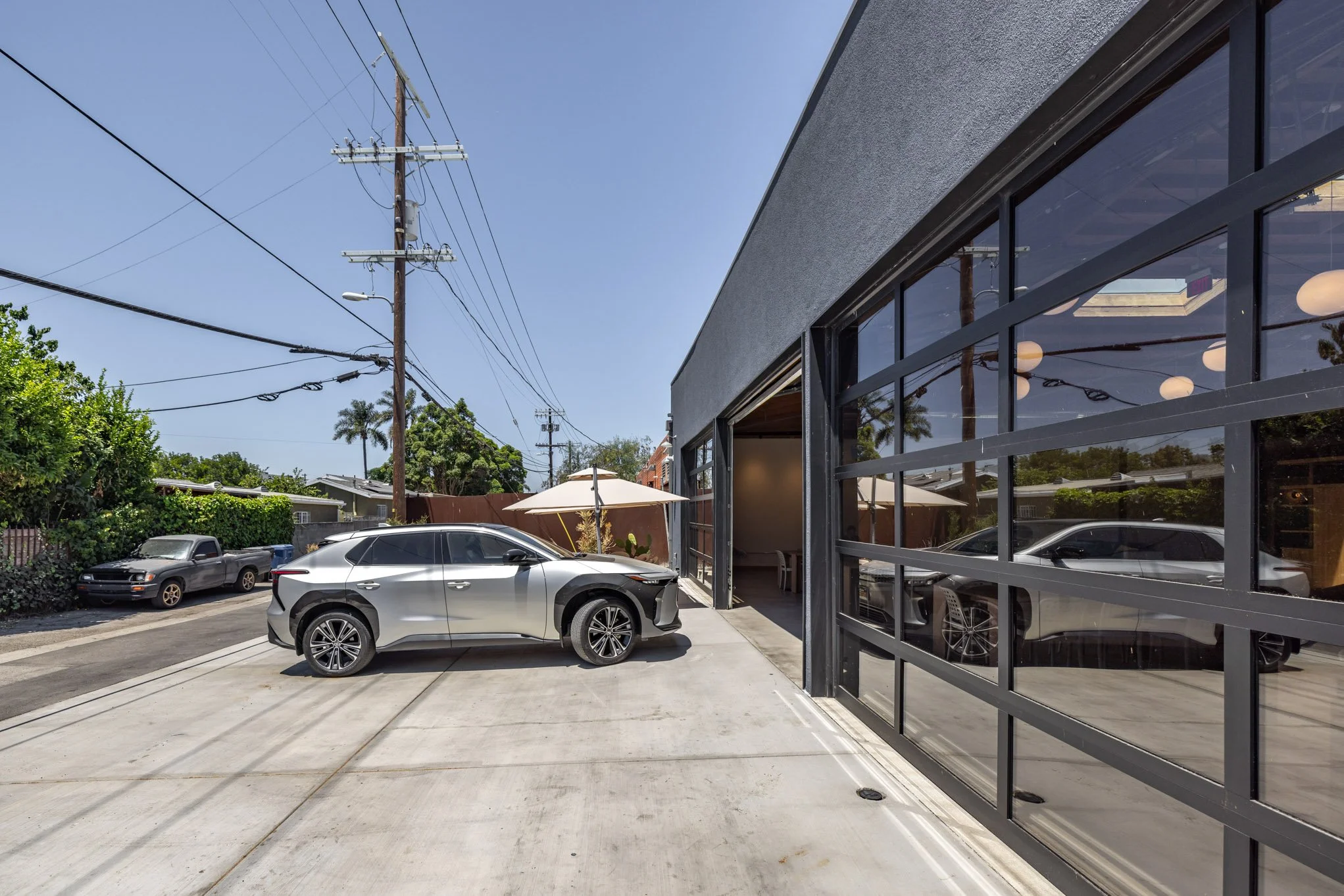 Silver SUV parked outside modern building with large glass windows and an open garage, sunny day, blue sky, power lines overhead, some trees and an umbrella visible in the background.