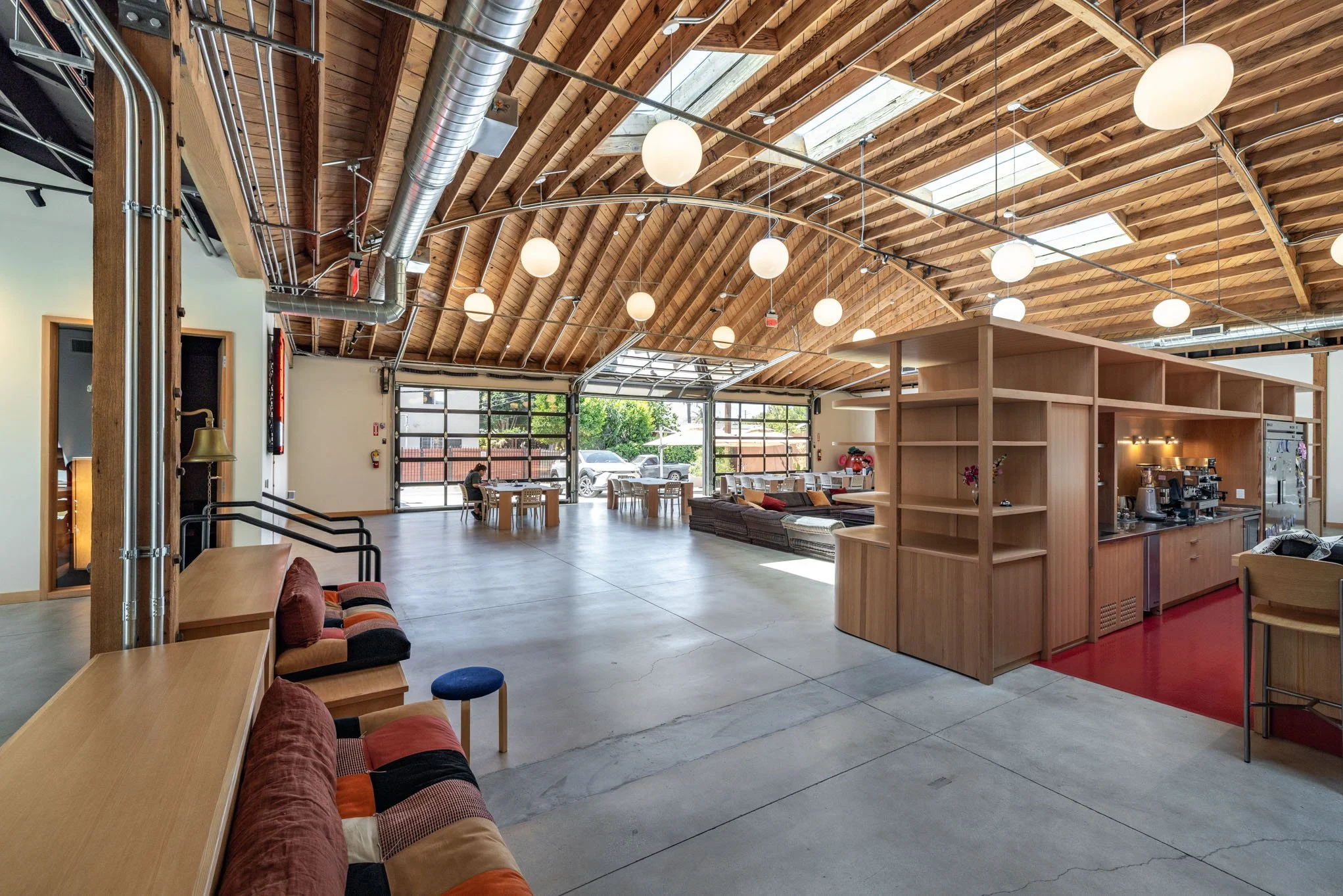 Spacious, modern cafe or community space with wooden vaulted ceiling, large glass garage-style doors, seating area, and a cafe counter with coffee machines.