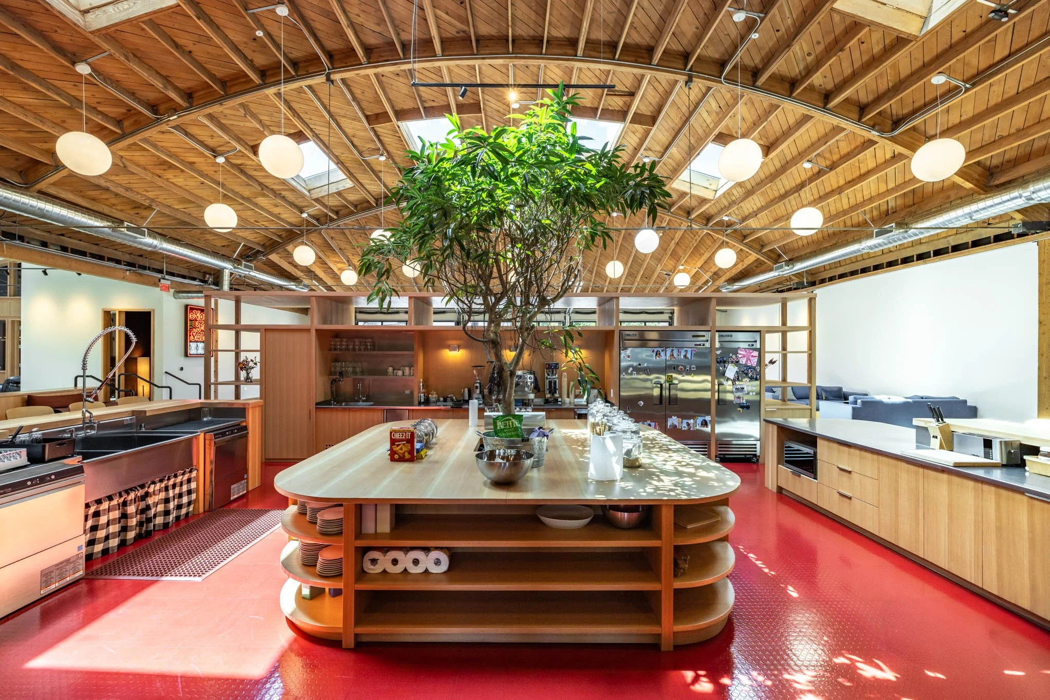 Modern kitchen with a large central island, wooden cabinetry, stainless steel appliances, and a large plant indoors under a wooden vaulted ceiling with large skylights and hanging lights.