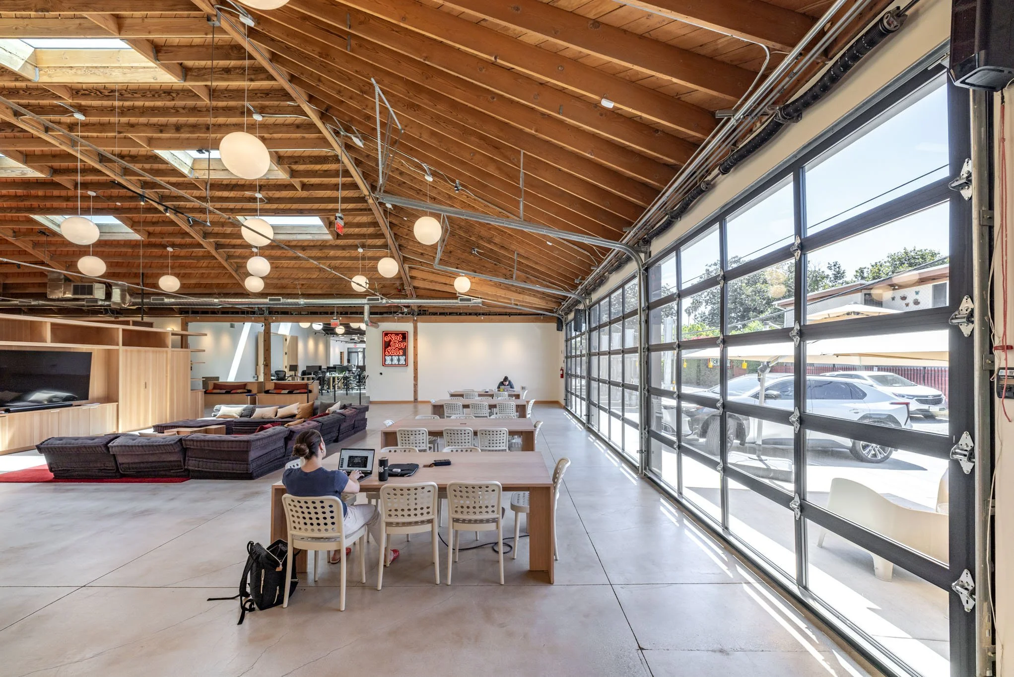 Interior of a spacious, modern office or coworking space with a high wooden ceiling, large glass garage-style doors, and seating areas including tables and couches. There are a few people working at desks and the space is well-lit with natural light.
