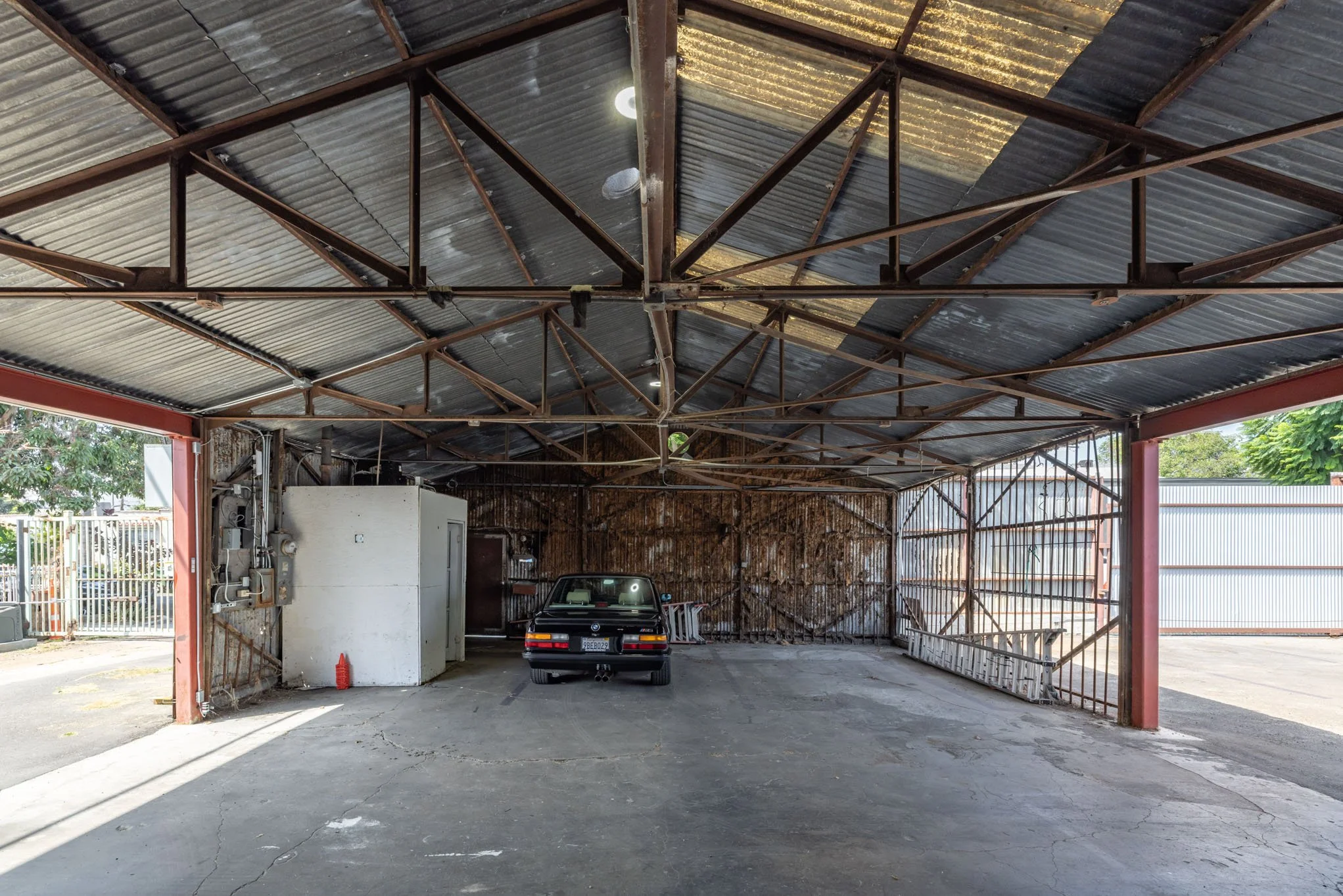 Empty parking garage with a single black car parked inside, metal roof and open sides, some electrical equipment on the left wall, and a cracked concrete floor.