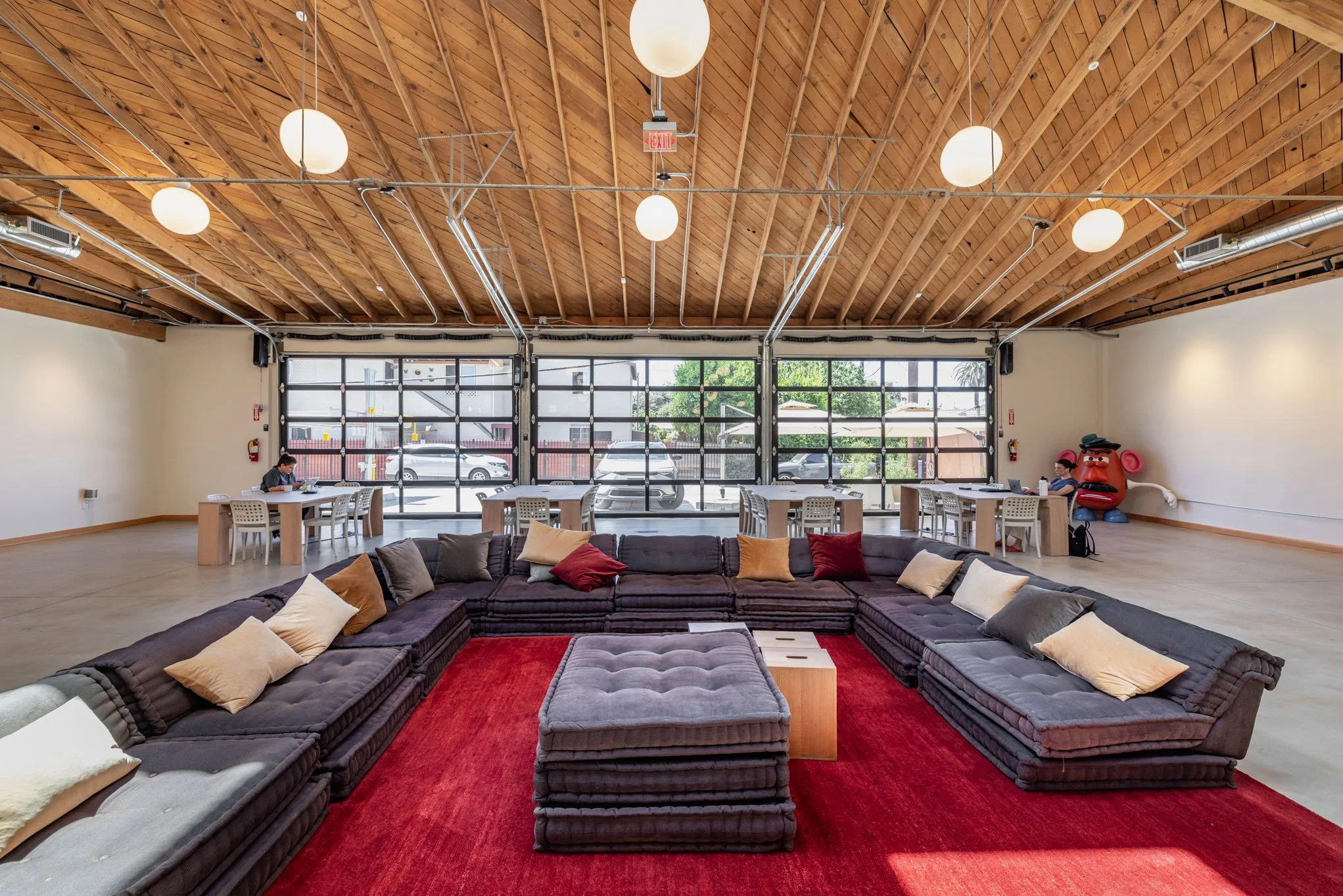 Spacious lounge area with a large sectional sofa, colorful pillows, and a red rug in the center. In the background, there are tables and chairs along a glass garage-style door, with a view outside and two people sitting at the tables. The ceiling is 
