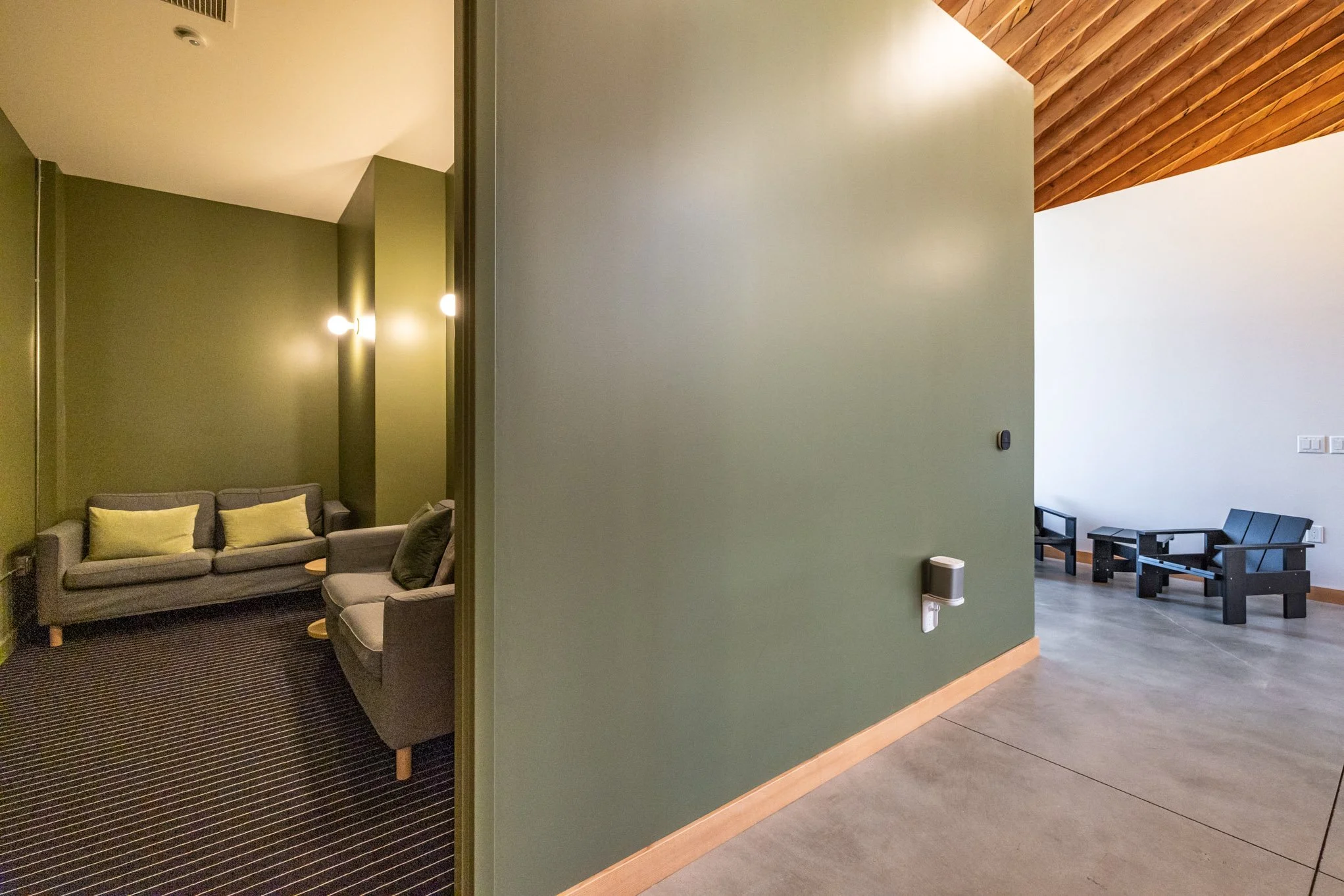 Interior view of a room with a green accent wall on the left, leading to a sitting area with gray couches and green cushions, and a corner with black chairs and a white wall with multiple light switches on the right.