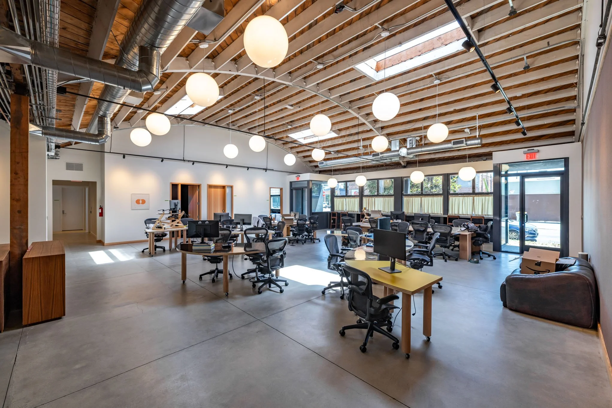 Open office space with multiple desks and black chairs, large windows along the back wall, white hanging spherical lights, concrete floor, exposed wooden beams and ceiling ducts, and a glass entrance door.