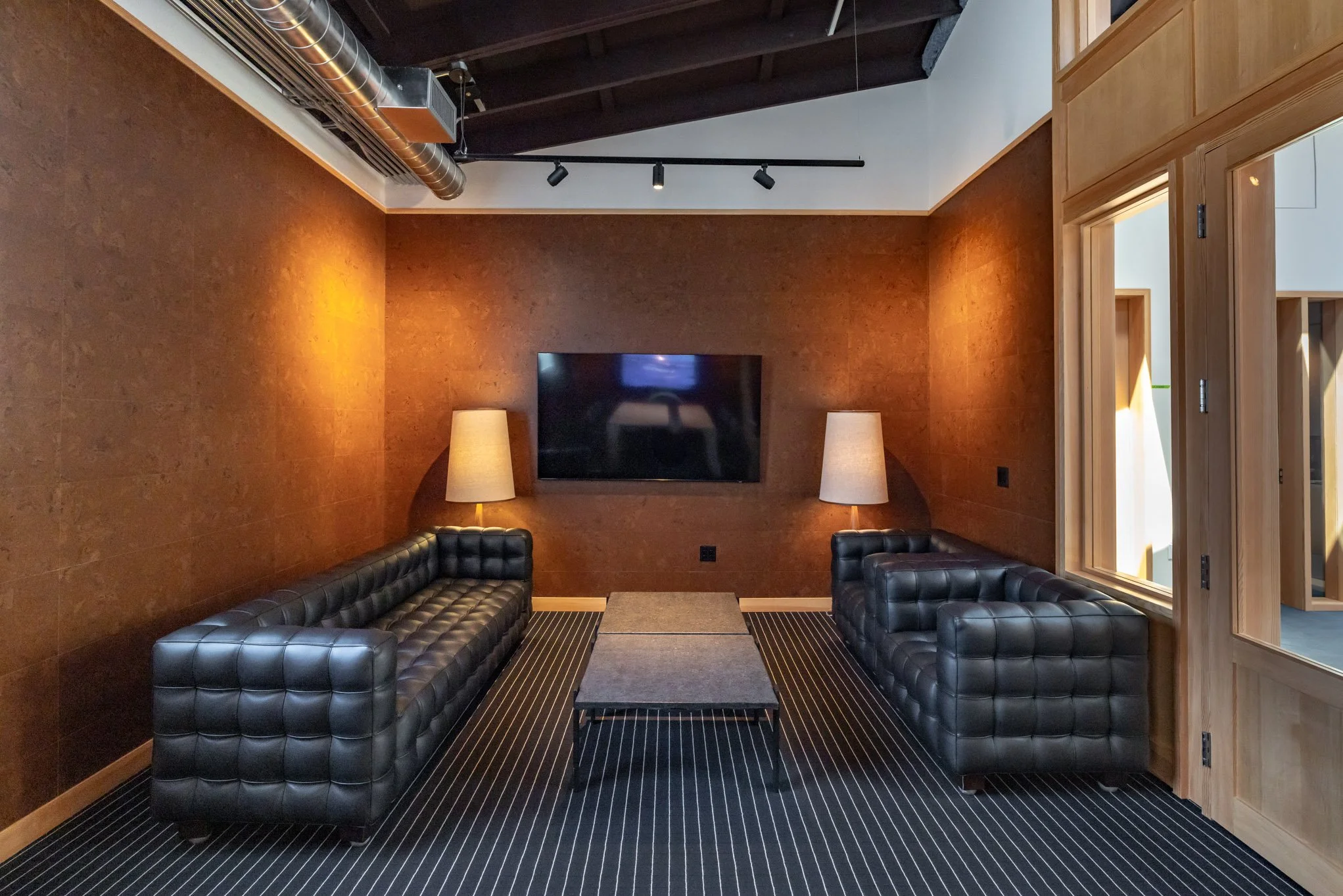 Modern lounge with two black leather tufted sofas, two white lamps, a flat-screen TV on a brown wall, a striped carpet, and wooden-framed windows and doors.
