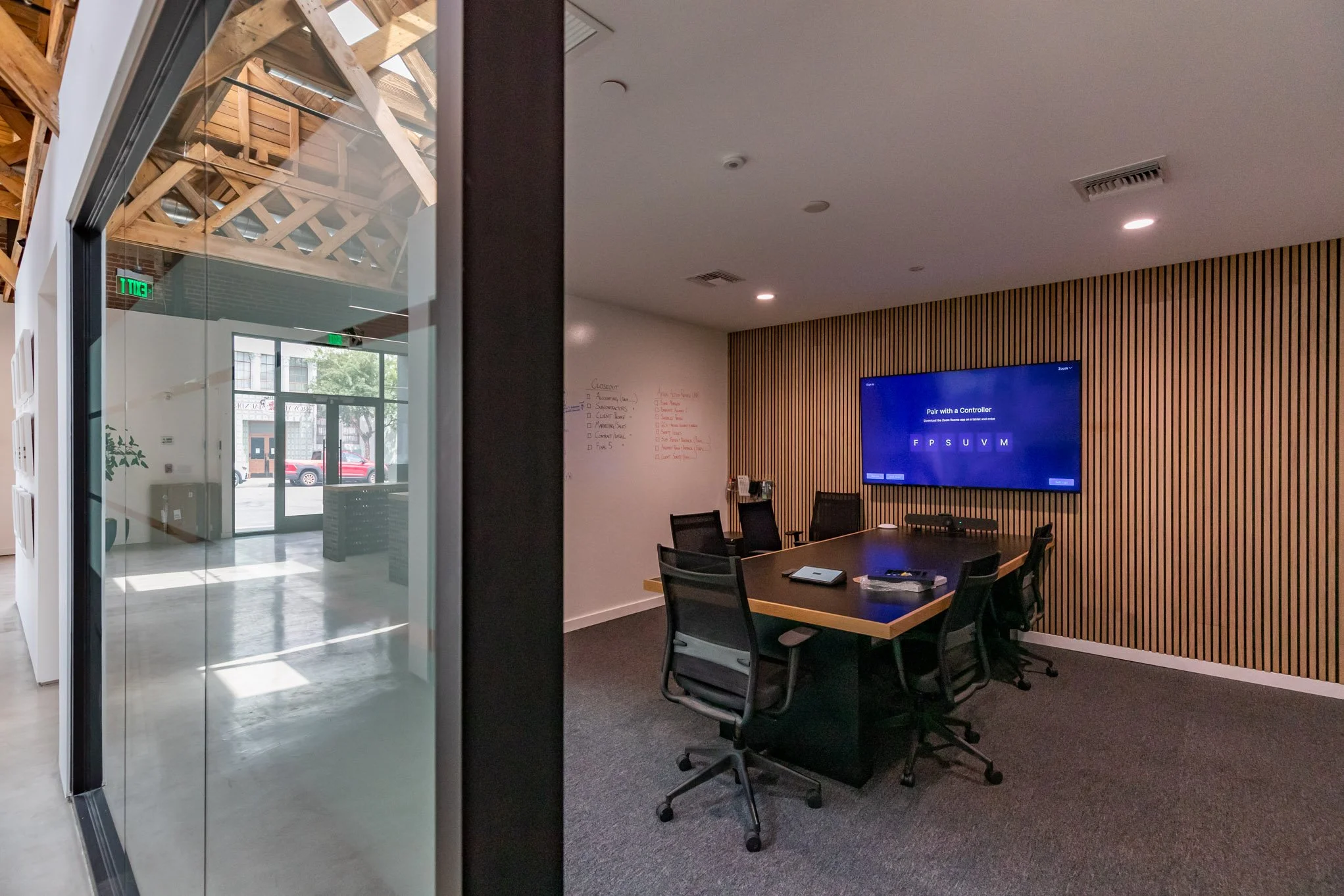 View of a modern conference room with a large table and six chairs, a wall-mounted TV screen displaying a task, and a whiteboard with notes inside a corporate office.