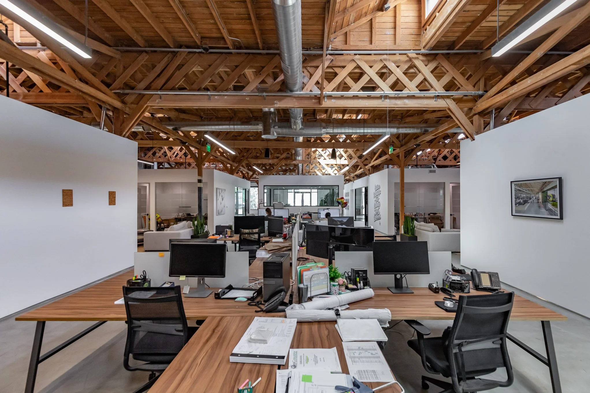 Open-plan office with wooden ceiling beams, desks with computers, office supplies, and chairs, white walls with artwork, and a large window in the background.