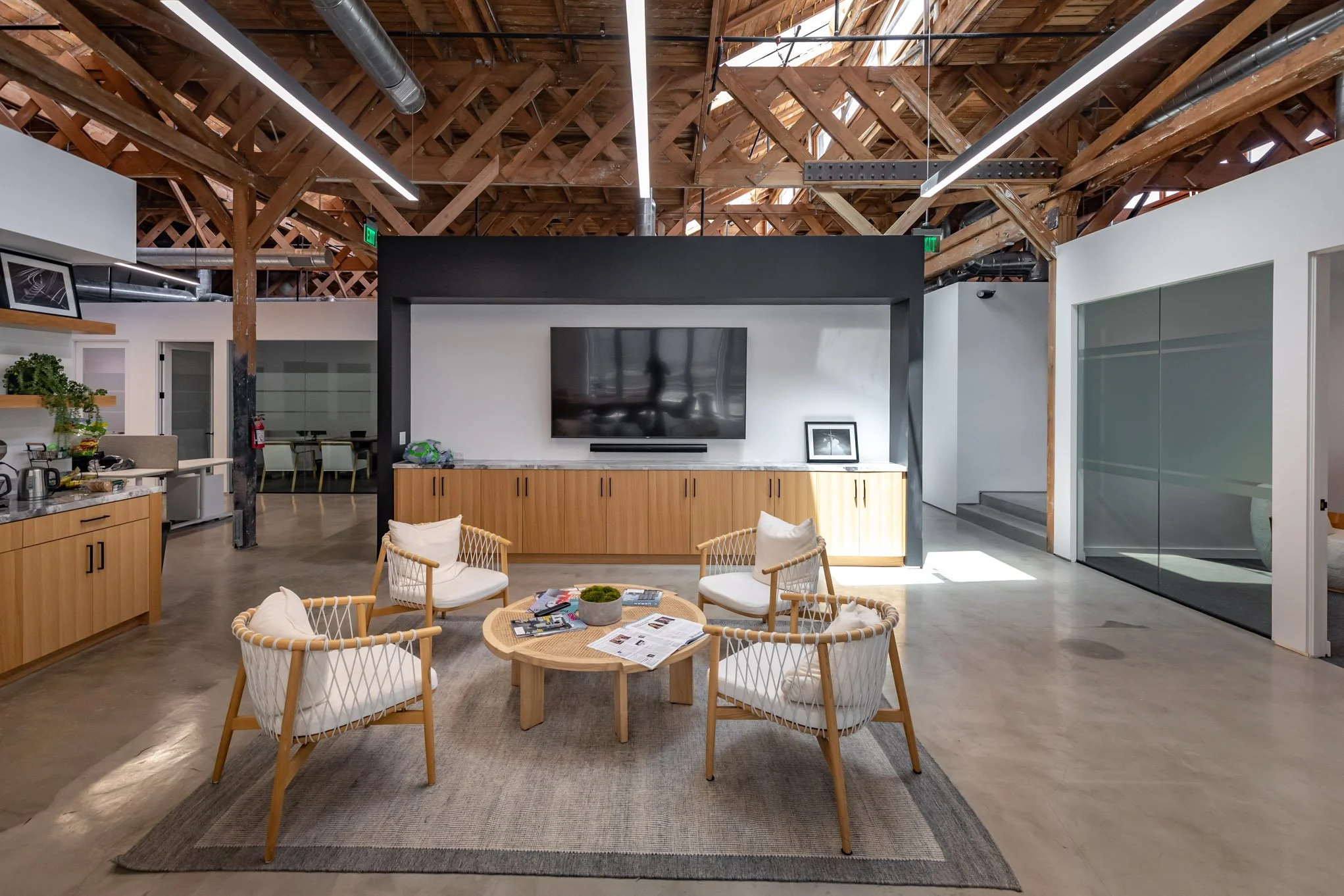 Modern lobby or lounge area with wooden beams and ceiling, featuring a central round table with magazines, four woven chairs with white cushions, a flat-screen TV on a black wall, a wooden cabinet below, and a gray and beige rug on polished concrete 