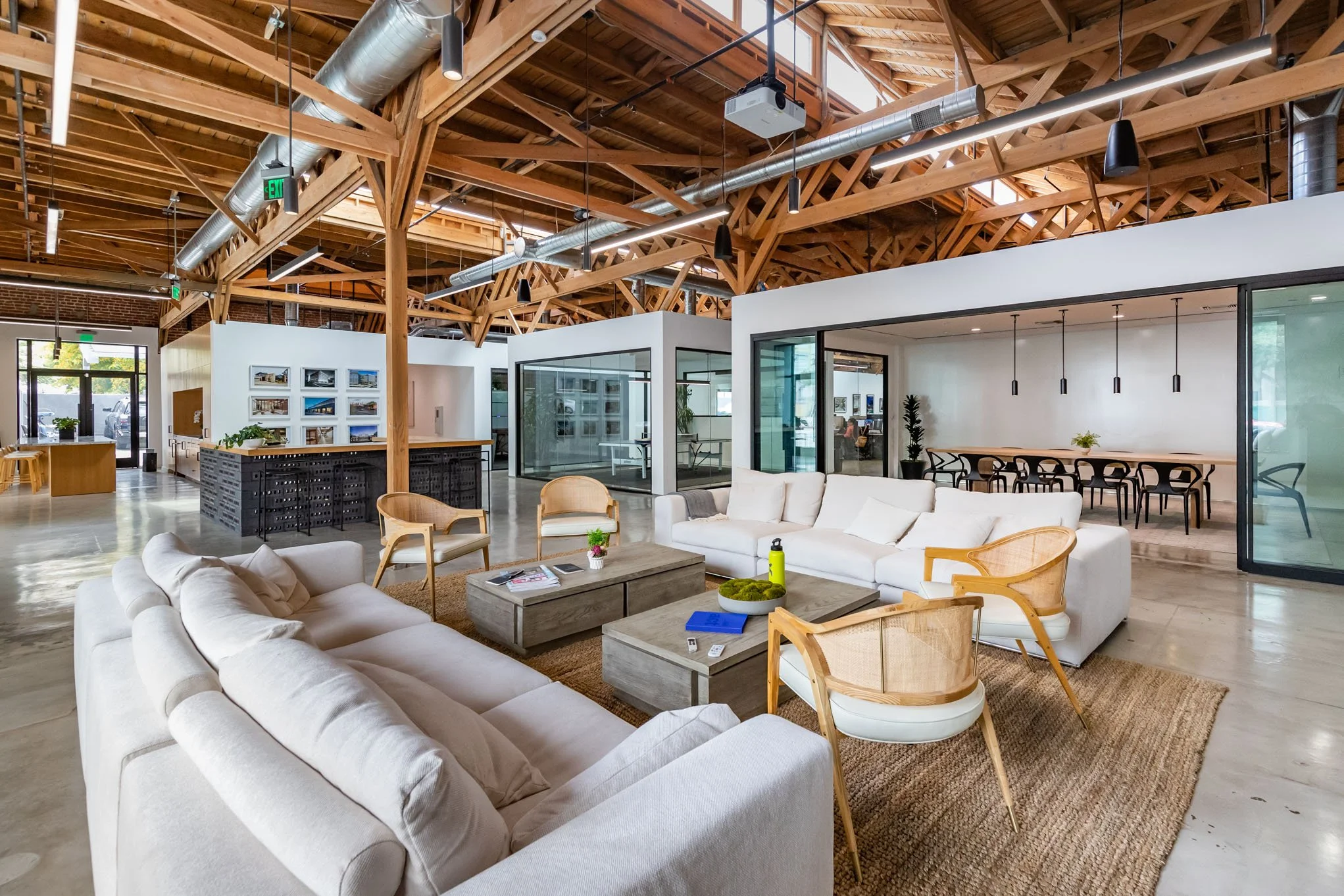 Modern open-plan office lounge with white sofas, wooden chairs, and a coffee table, featuring exposed wooden beam ceilings and glass-walled meeting rooms.