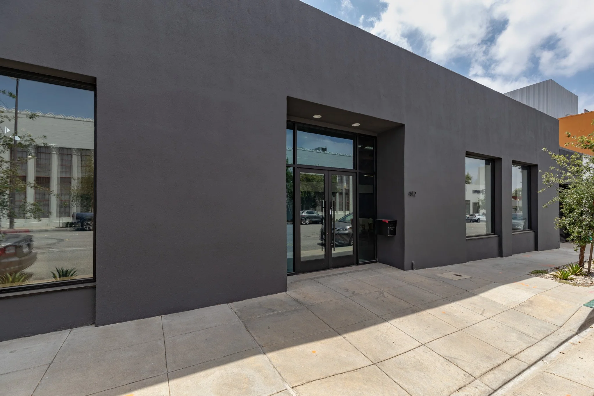 Modern gray building with large glass door and windows, sidewalk in front, a small tree on the right, and parked cars reflected in the windows.
