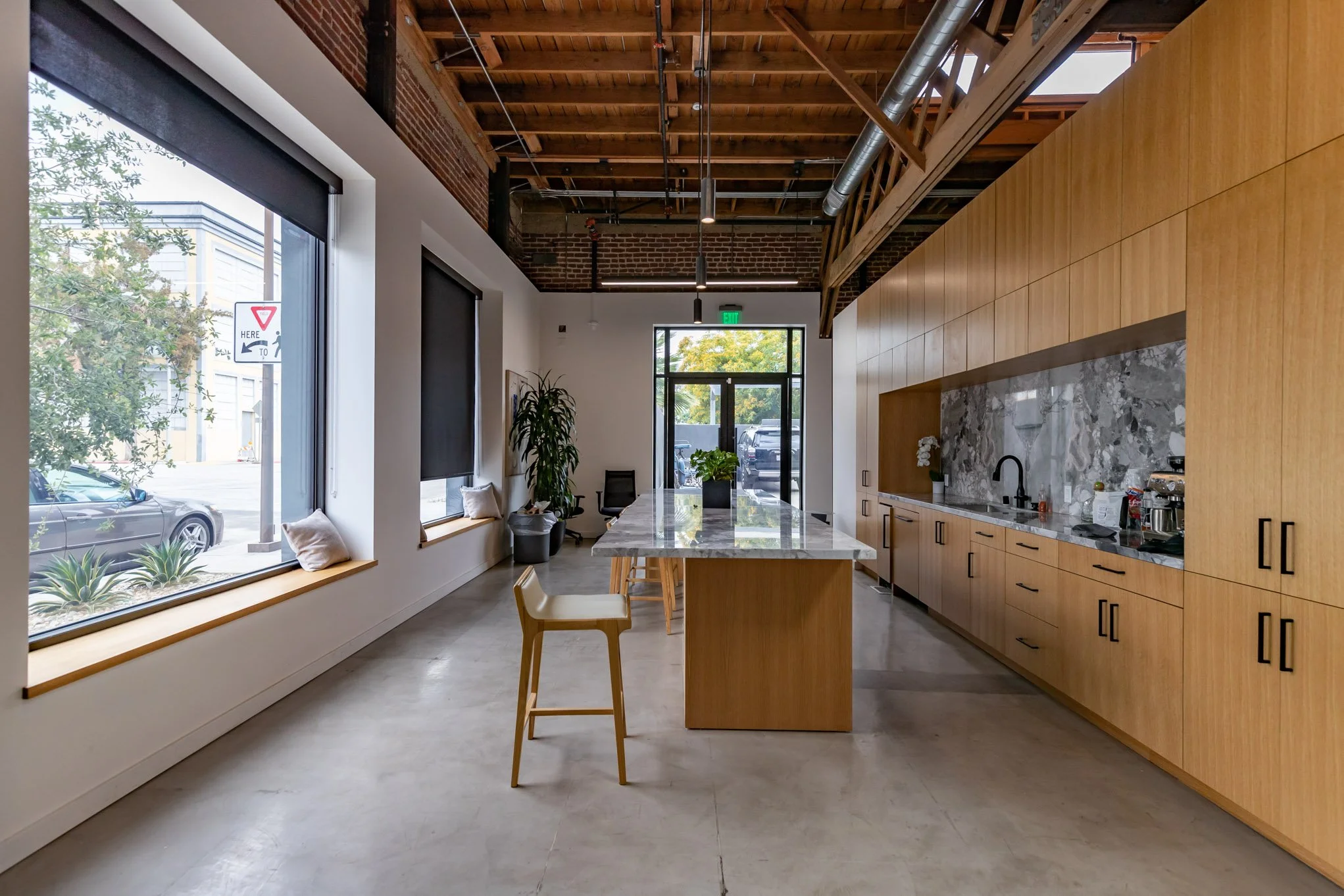 Modern kitchen with wooden cabinets, marble backsplash, island with a flower pot, large windows with black blinds, and a polished concrete floor in an industrial-style space.