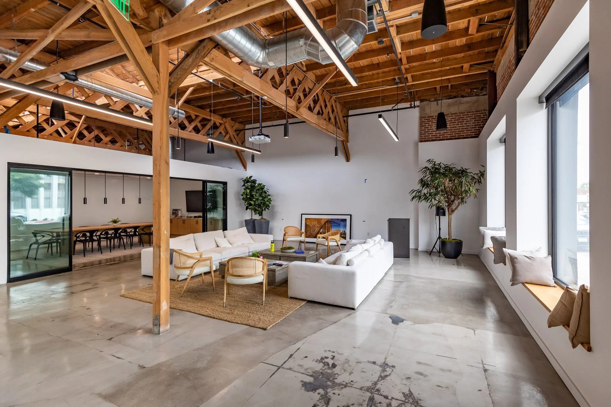 Spacious modern living room with high wooden ceiling, exposed ductwork, large windows with window seats, white sofas, wooden chairs, potted plants, and minimalist decor.