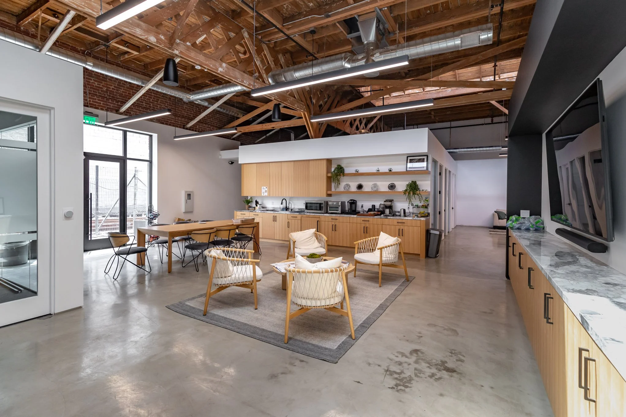 Open office kitchen with wooden cabinets, a small seating area with four chairs around a circular table, and a long table with chairs near a large window and glass door. Exposed wooden ceiling beams and ductwork are visible.