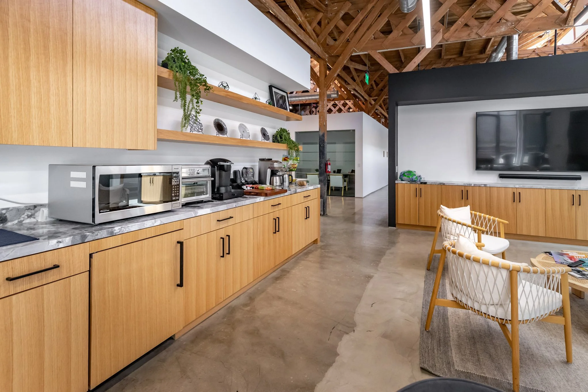 Modern kitchen and lounge area with wooden cabinets, open shelving, kitchen appliances, a large flat-screen TV, and seating with a glass vaulted ceiling.