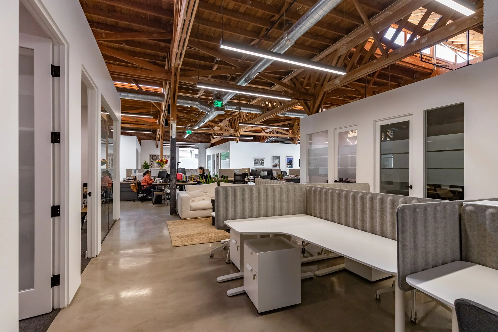 Open-concept office with exposed wooden ceiling beams, modern lighting, glass-walled meeting rooms, work desks with computers, and a few employees working.