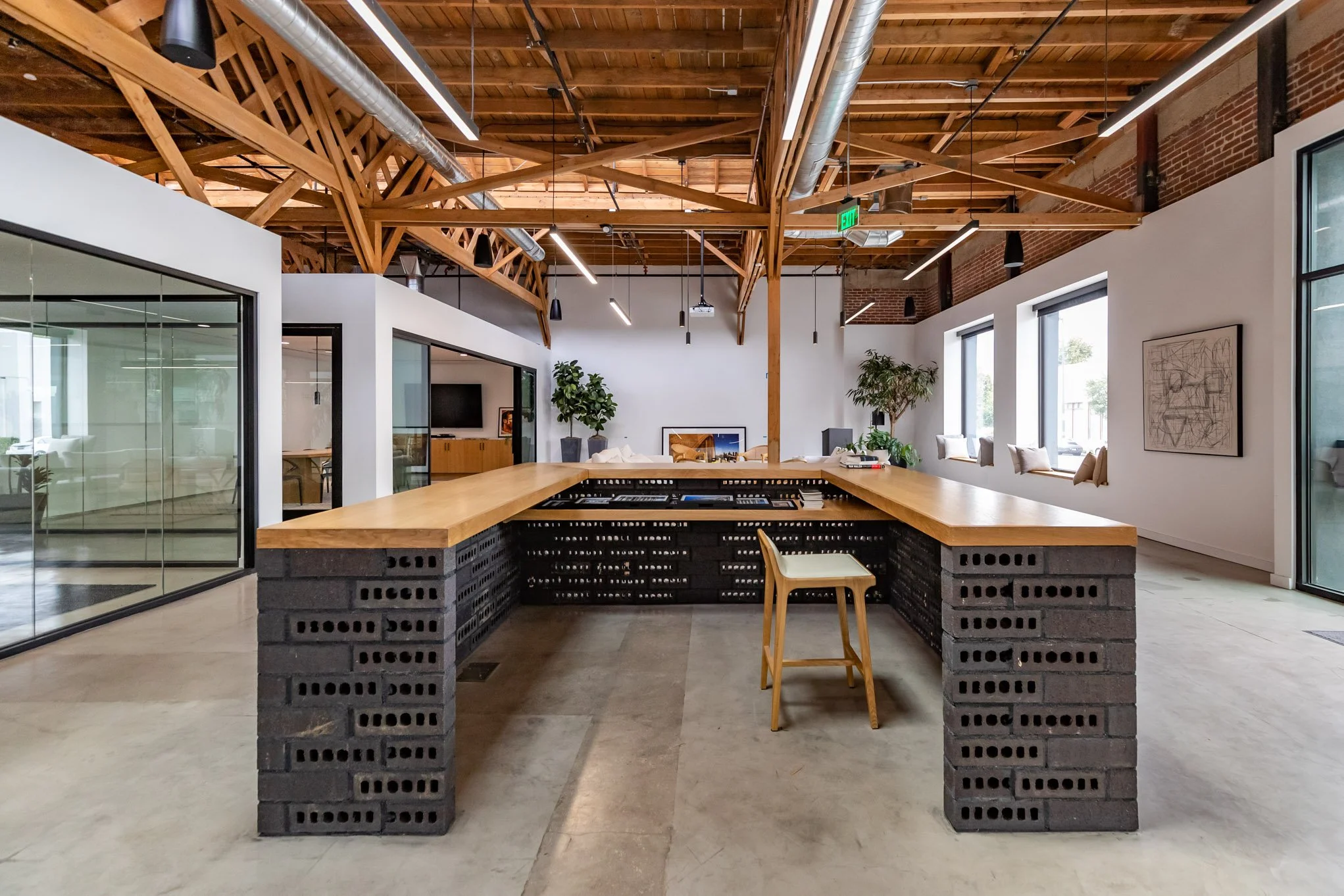 Modern open-concept office space with a wooden ceiling, exposed beams, and large windows, featuring a central bar-height table made of wood and black concrete blocks, with a single chair, surrounded by plants and artwork.
