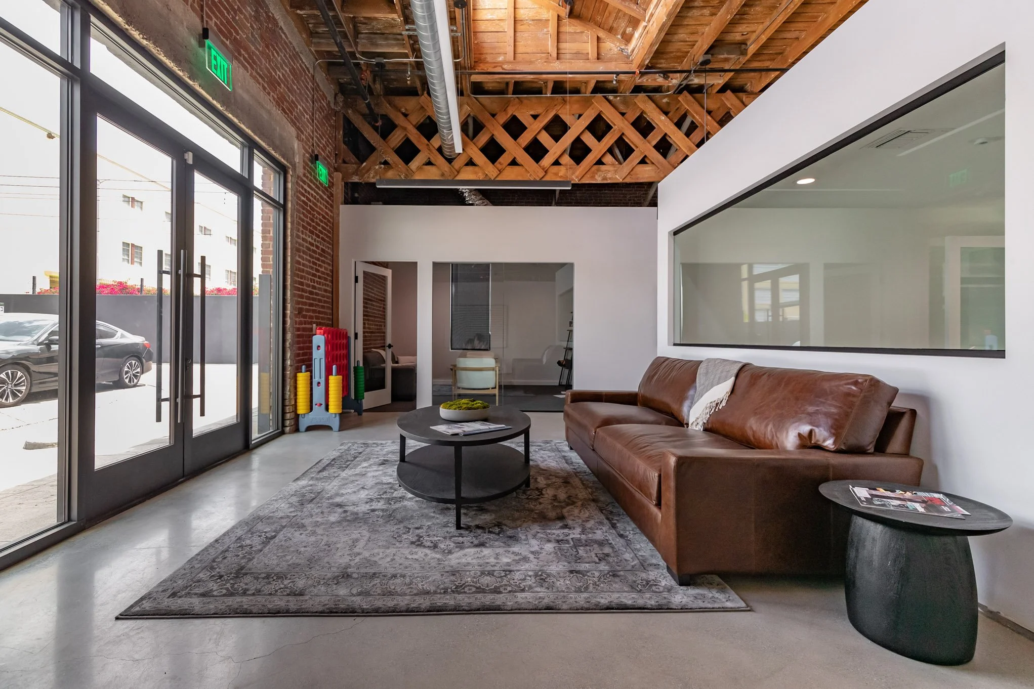 Modern lobby with brown leather sofa, black coffee table, side table with magazines, large windowed wall, exposed brick wall, and decorative ceiling beams.