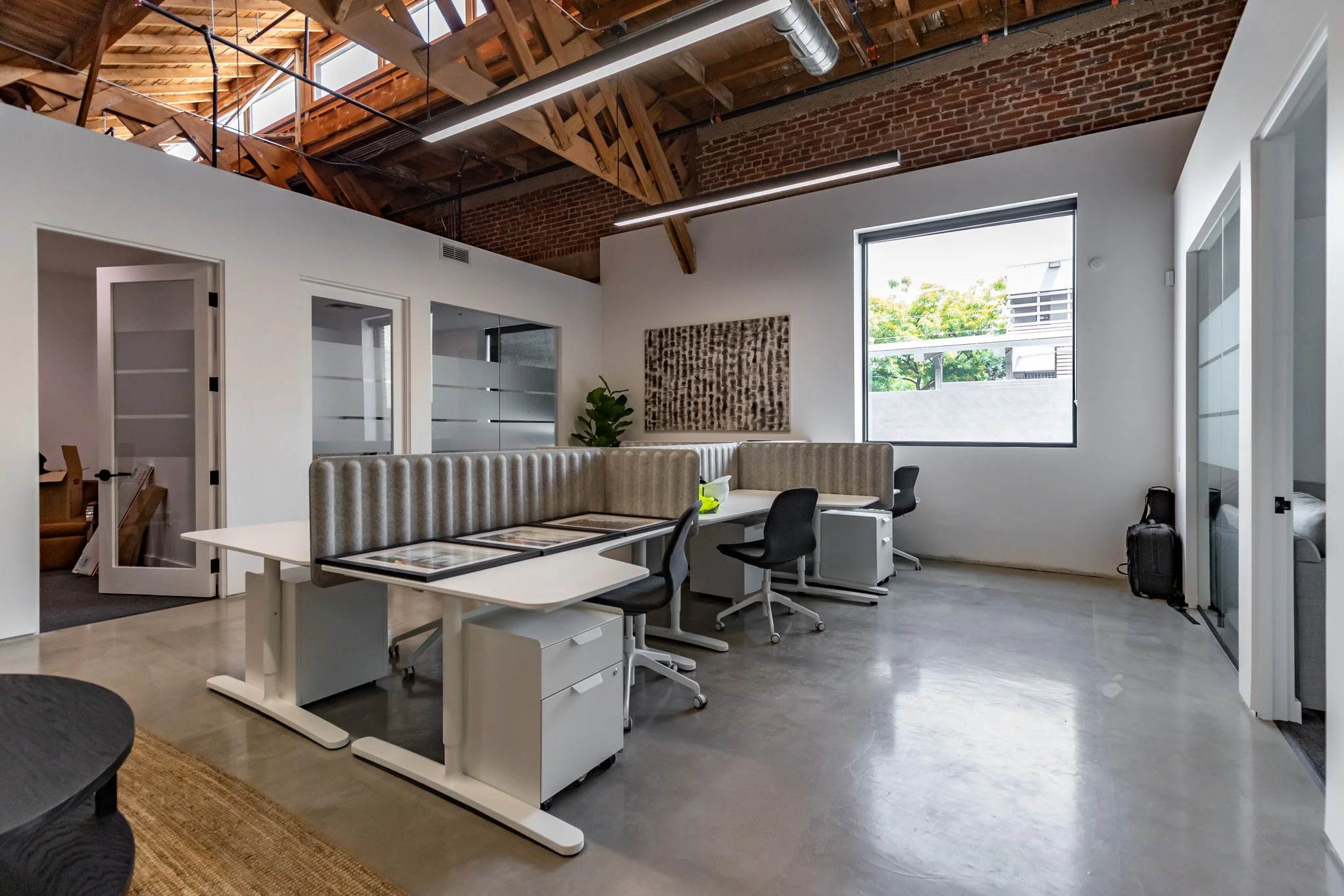 Office space with white walls, high wooden ceiling with exposed beams, conveyor ductwork, and a large window. Desks with black chairs and beige upholstered privacy panels, and a potted plant near the wall.