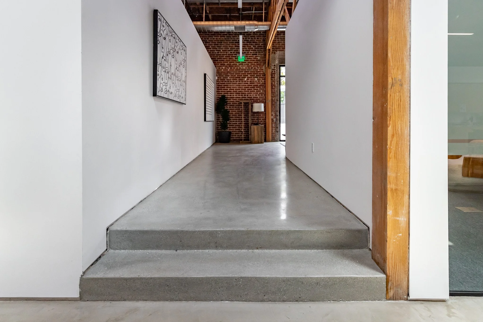 Modern interior hallway with exposed brick wall, white walls, concrete floor, wooden accents, and minimal decor, including a potted plant, a lamp on a wooden stand, and wall art.