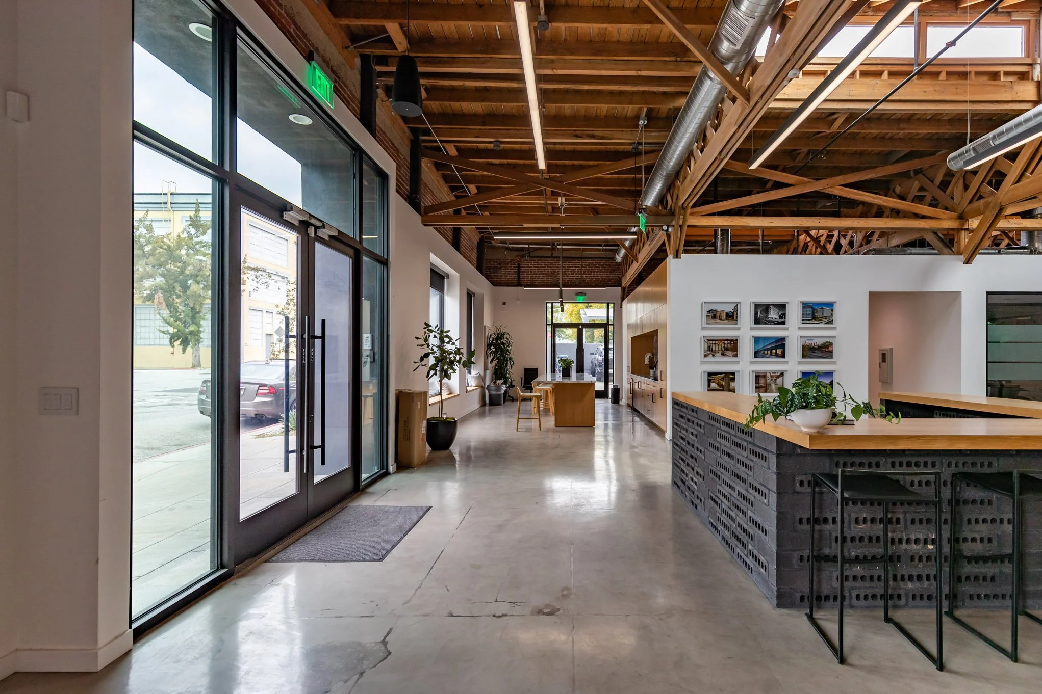 Interior of a modern office space with high ceilings, exposed wooden beams, large windows, a reception counter with a potted plant, and seating area near the far end.