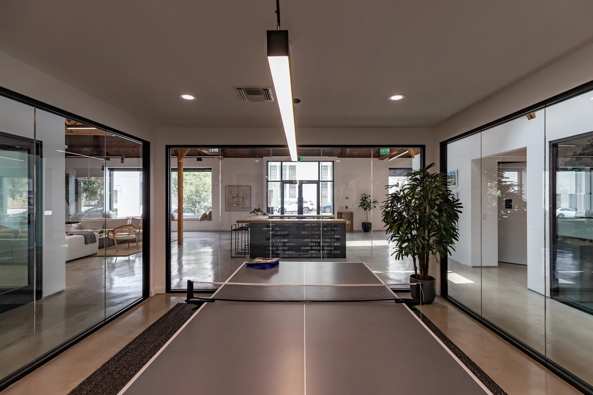 Indoor recreational room with table tennis table, potted plant, large glass windows, and seating area with sofas.
