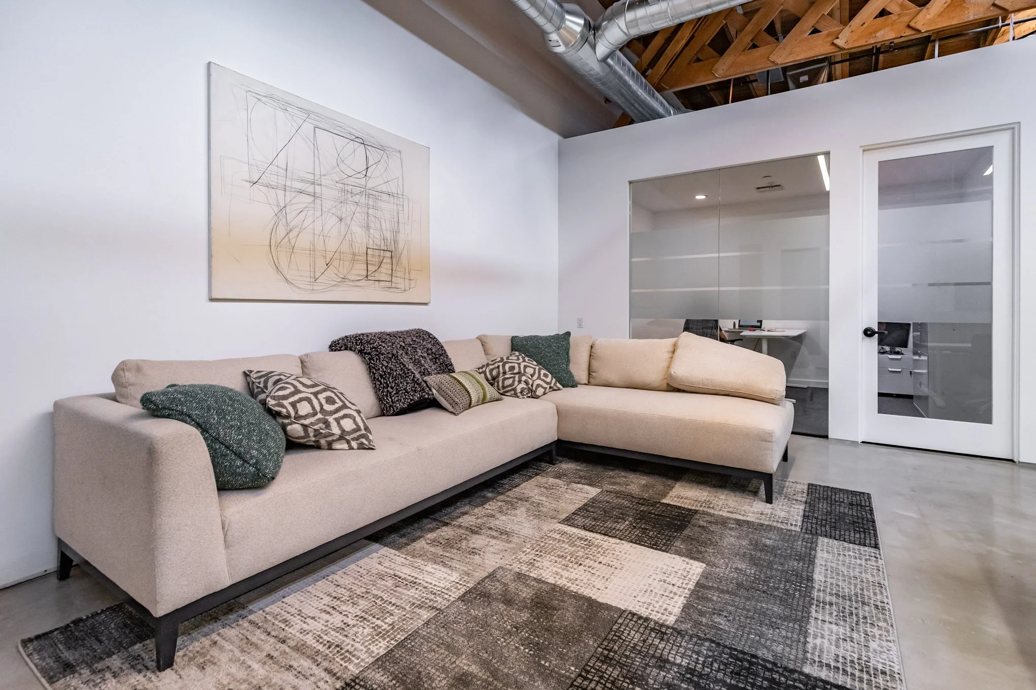 Modern living room with beige sectional sofa, patterned pillows, abstract artwork on white wall, glass office door, exposed wooden ceiling beams, and industrial ductwork.