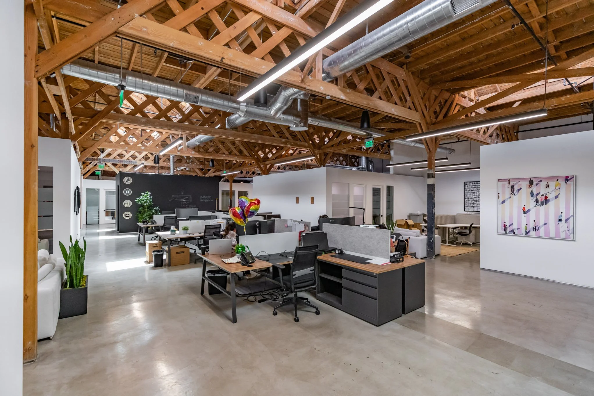 Open-plan modern office with wooden ceiling beams, exposed ductwork, and multiple workstations, some with computers and office supplies, along with colorful balloons, potted plants, and artworks on white walls.