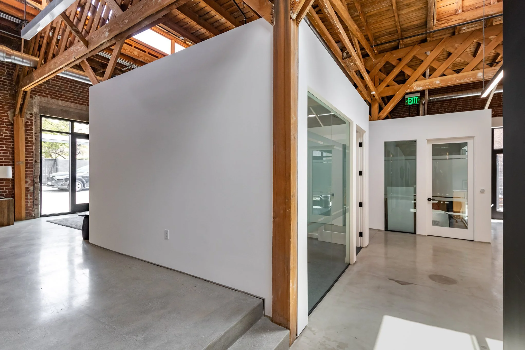 Interior of a modern office space with a white partition wall, glass doors, exposed wooden beams, and a concrete floor.