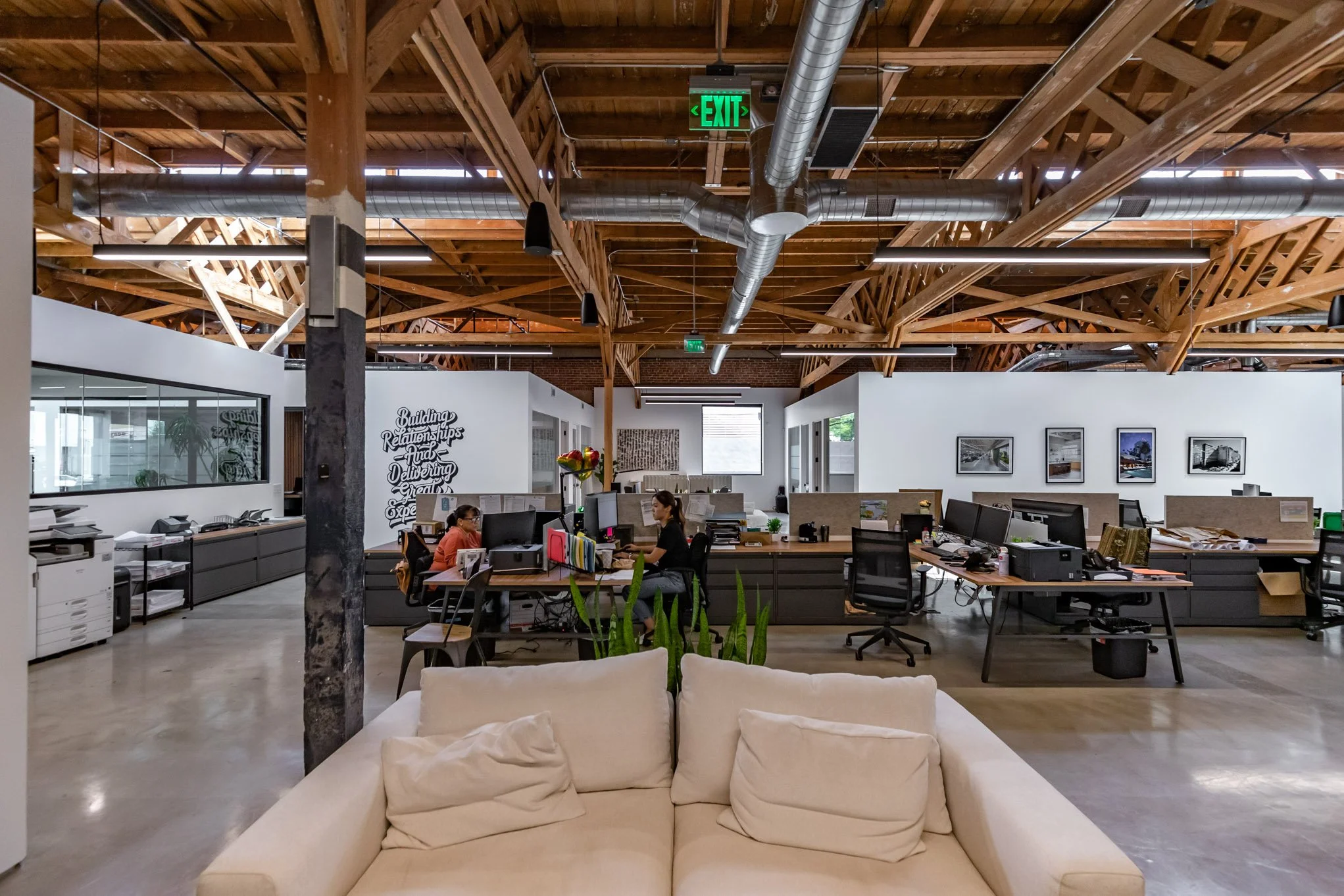 Open-plan office with exposed wooden beams, modern desks and chairs, a white sofa in the foreground, and artworks on the walls.