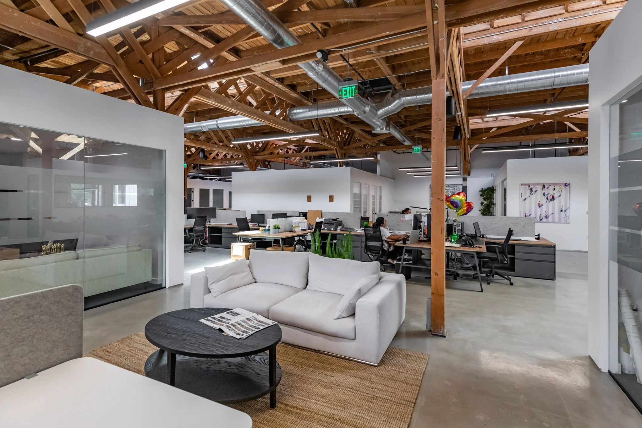 Open-plan modern office with a white sofa, round black coffee table, workstations with computers, wooden ceiling beams, and exposed ductwork.