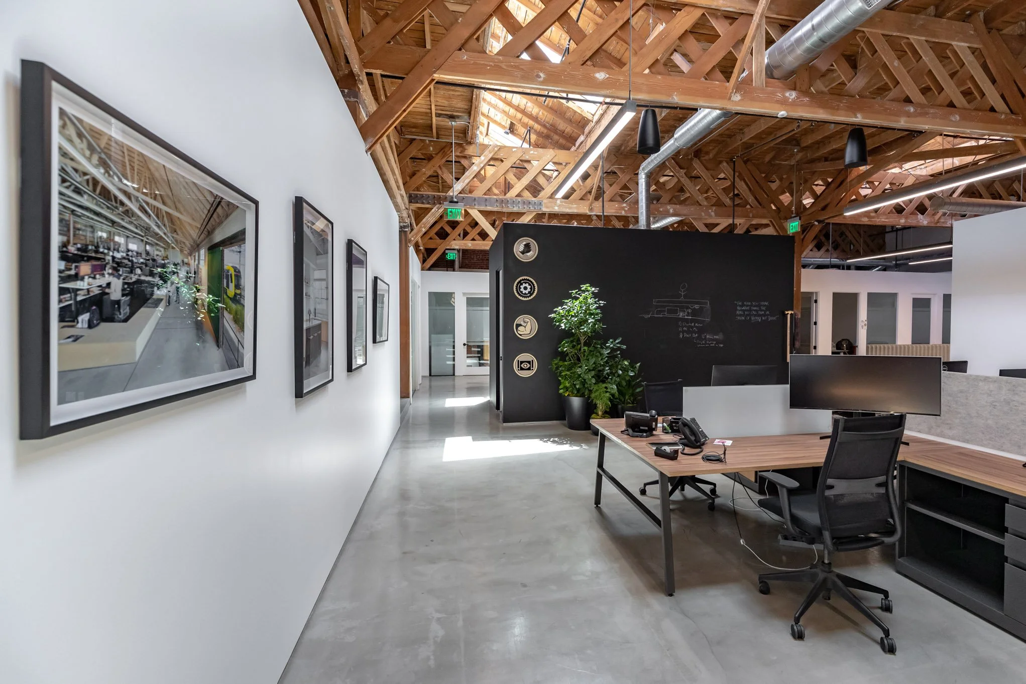 Modern office space with white walls, framed photos, a black chalkboard wall with sketches, a wooden desk with dual monitors, a black office chair, and exposed wooden ceiling beams.