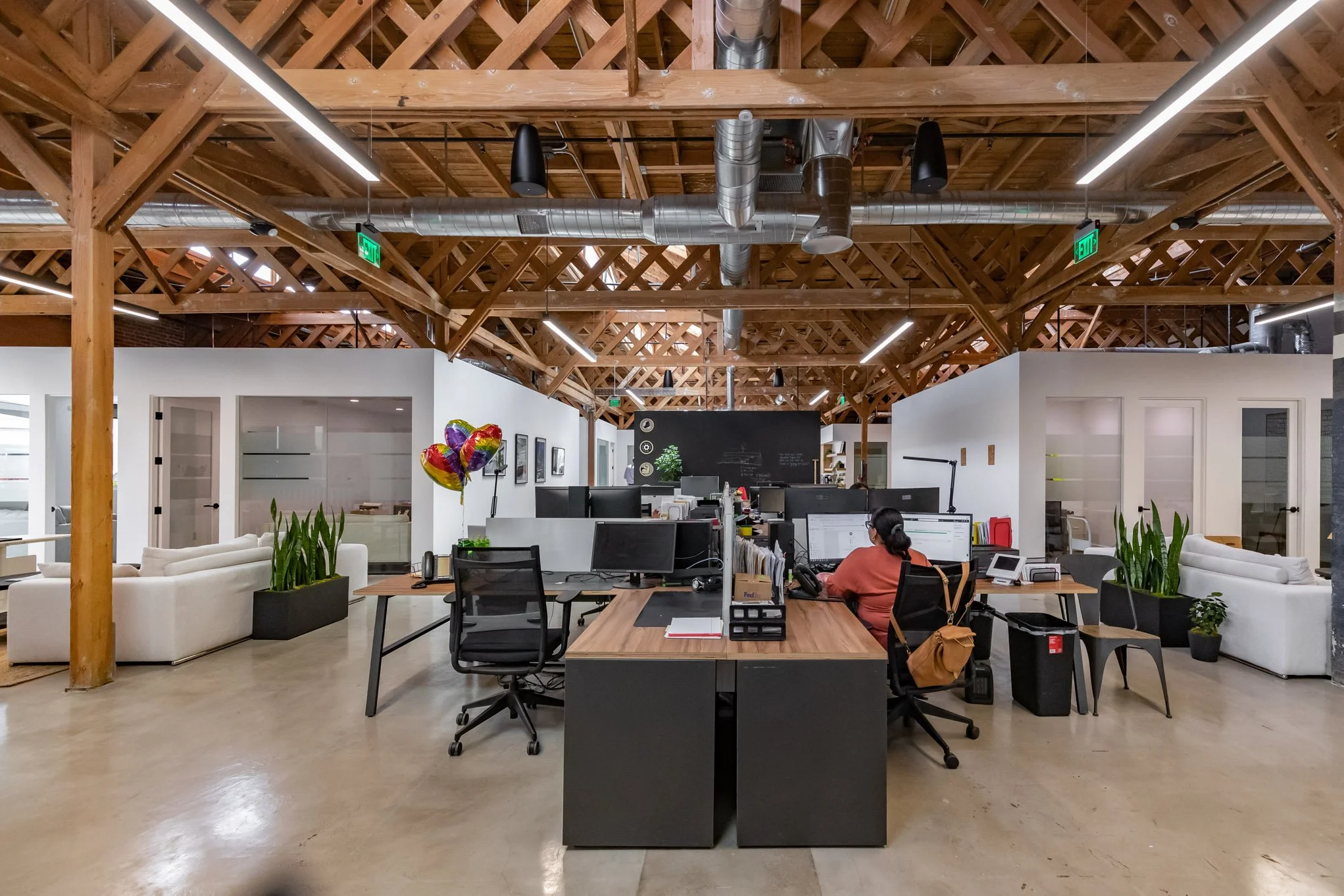 Open-plan office with wooden exposed beams ceiling, desks with computers, a woman working at a station, lounge area with white sofas, green plants, and balloons.