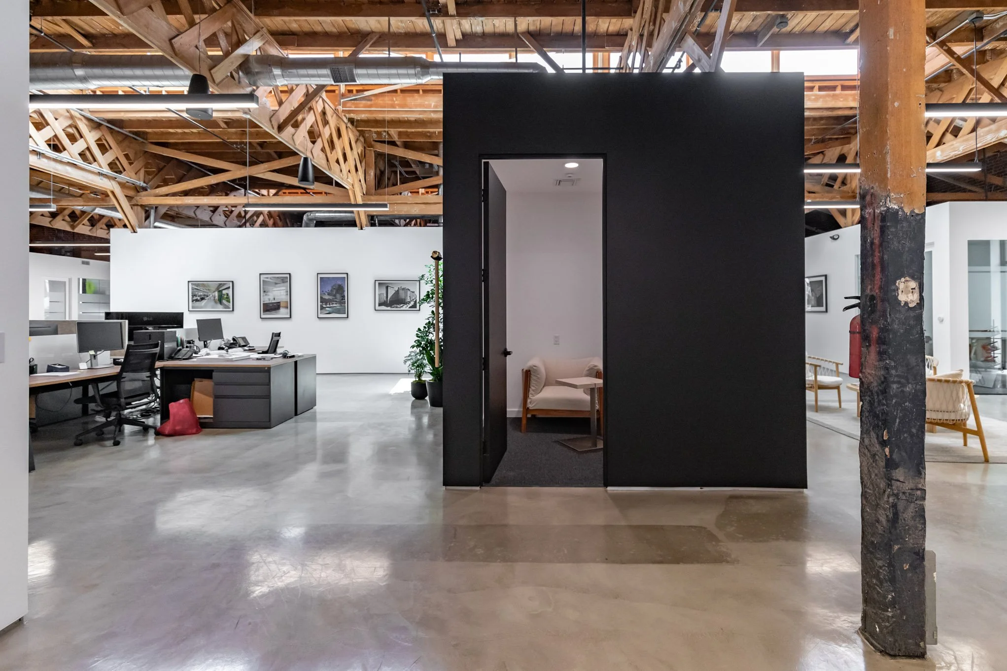 Open office space with exposed wooden ceiling beams, a black wall with a doorway, and a separate seating area with a beige loveseat and chairs.