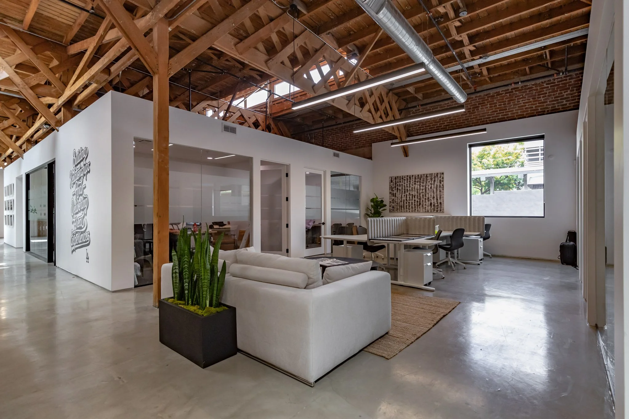 Modern office space with exposed wooden beams, white walls, a glass-enclosed meeting room, a white couch, a potted plant, and desks with chairs near a large window.