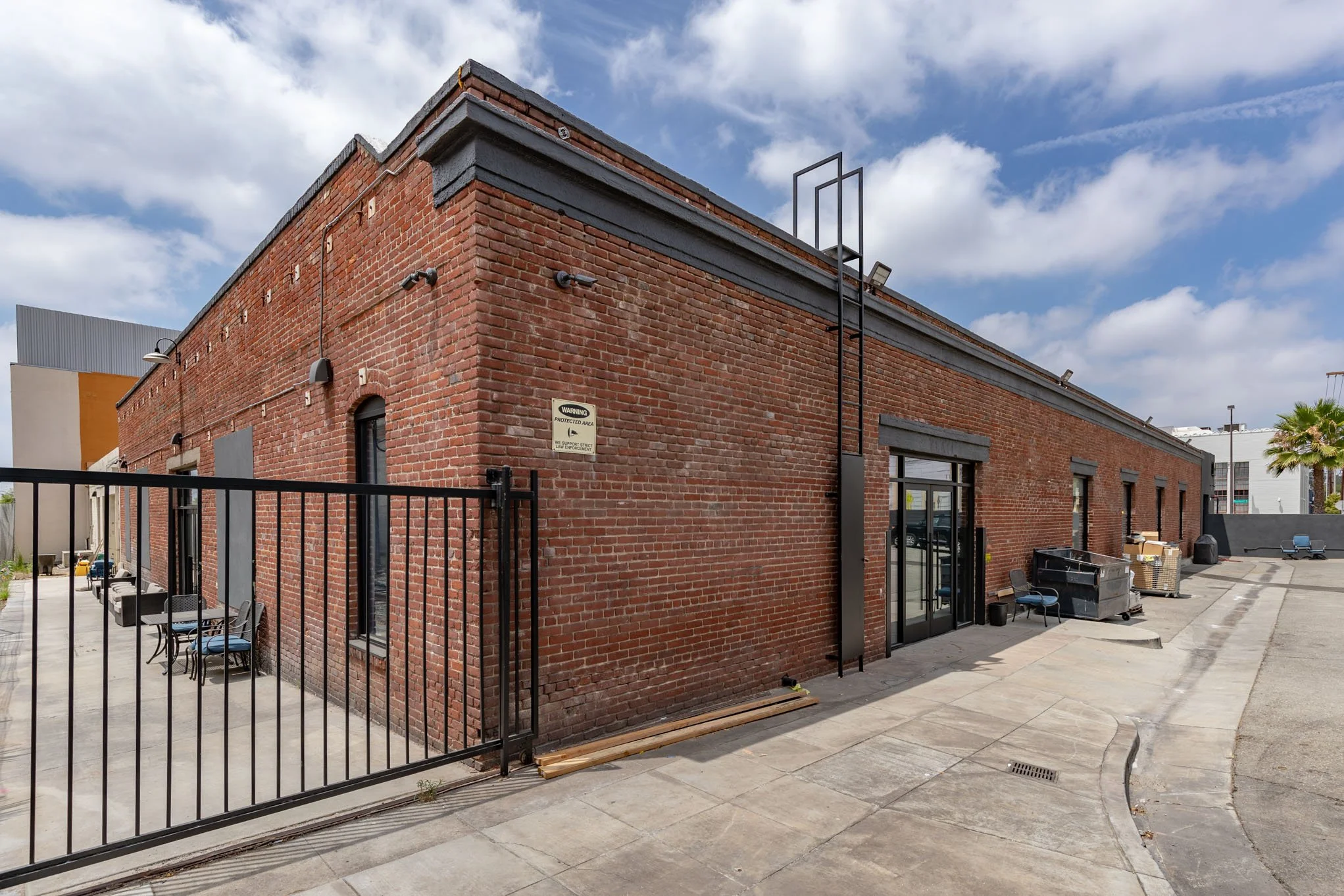 Red brick building with multiple windows, a black metal door, and outdoor chairs and tables on the patio. The sky is partly cloudy with blue sky visible. There's a black fence gate at the corner of the building and some construction materials on the 