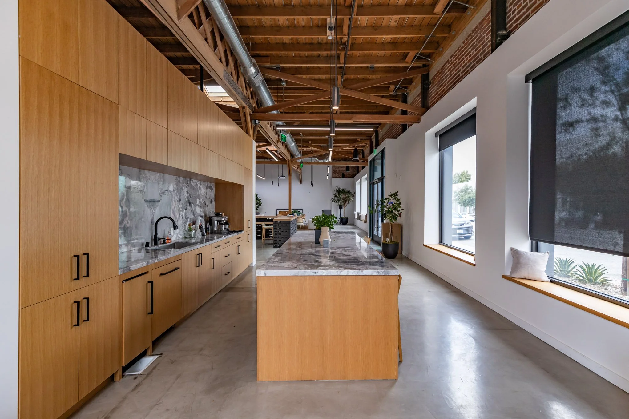 Modern industrial-style kitchen and lounge area with large windows, wooden beams, and minimalist decor.