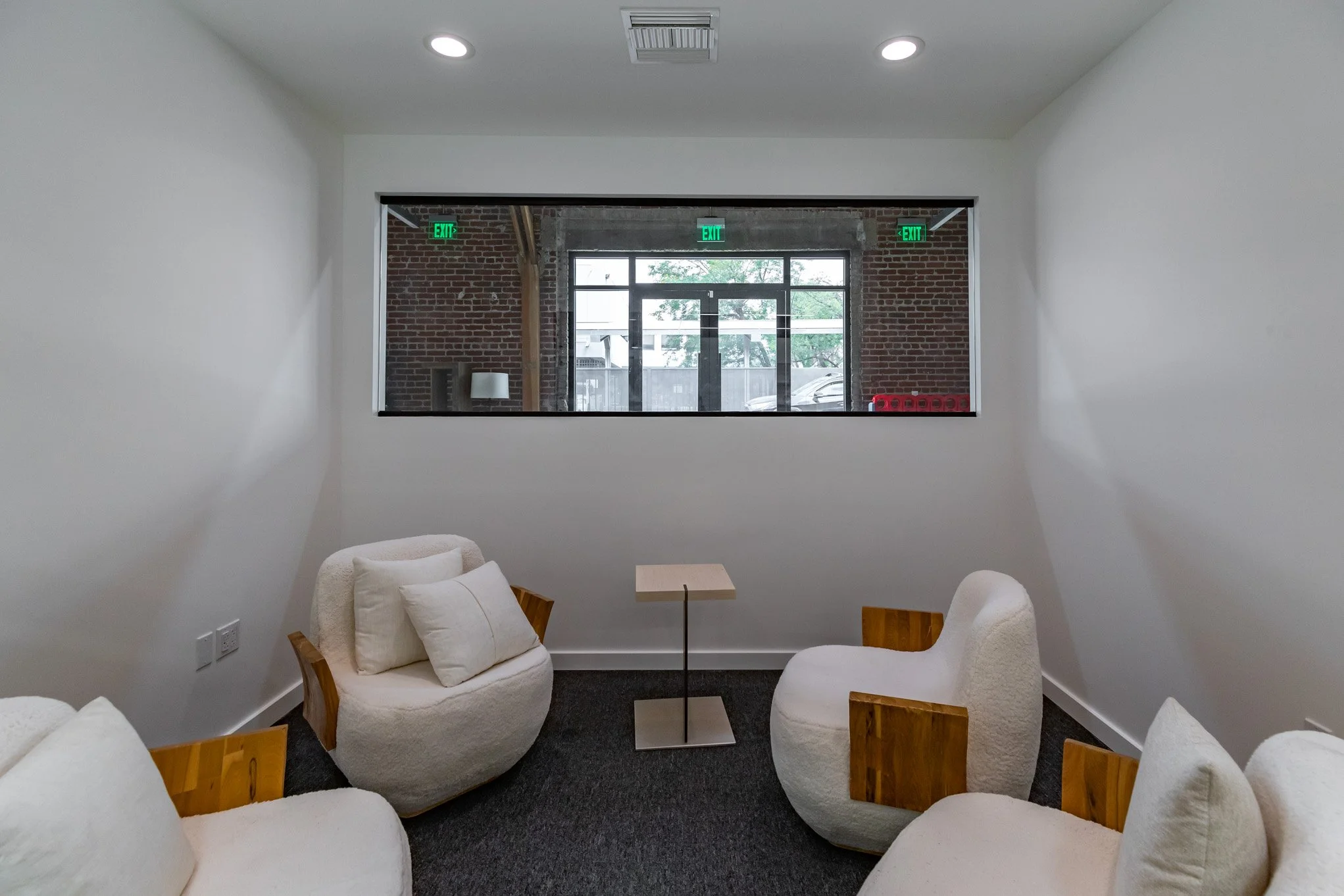 A small room with white walls and a large window looking outside. The room contains three rounded armchairs with wooden armrests, each with white cushions, and a small square table in the middle. Outside, there are brick walls and trees visible throu